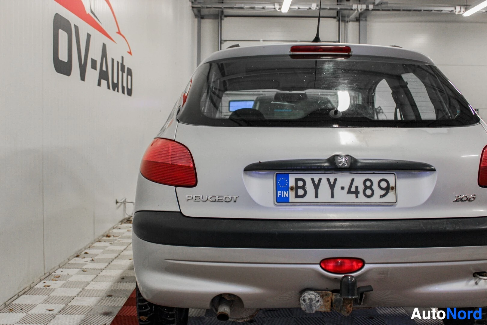 Takaa: Hopeisen Peugeot 206:n takaosa kuvattuna suoraan takaa. Rekisterikilvessä lukee. Autossa on vetokoukku