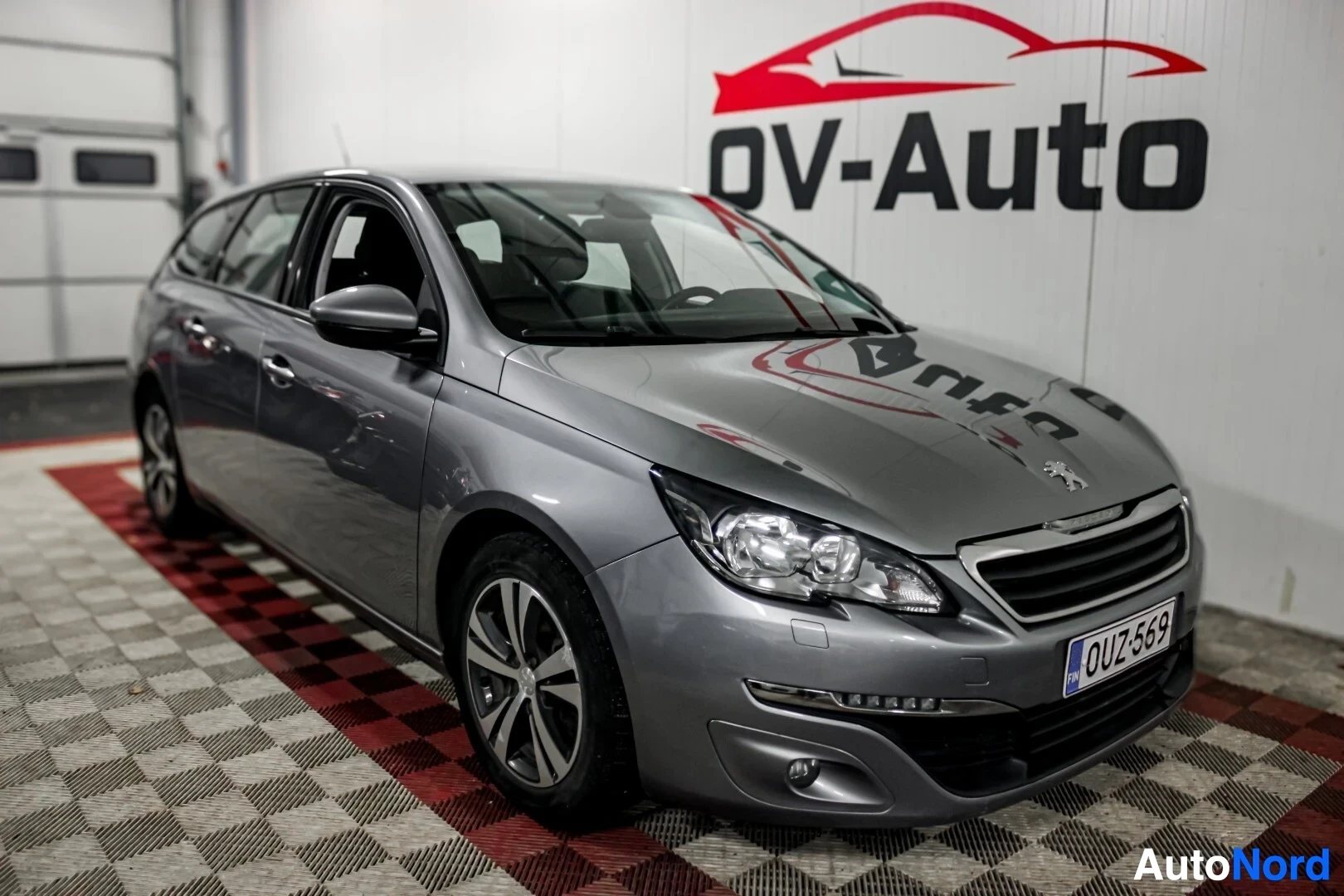 Etuviisto, nokka vasemmalle: Harmaa Peugeot 308 -farmariauto edestä ja vasemmalta, kuvattuna sisätiloissa ruudullisella lattialla ja OV-Auton logolla seinällä.