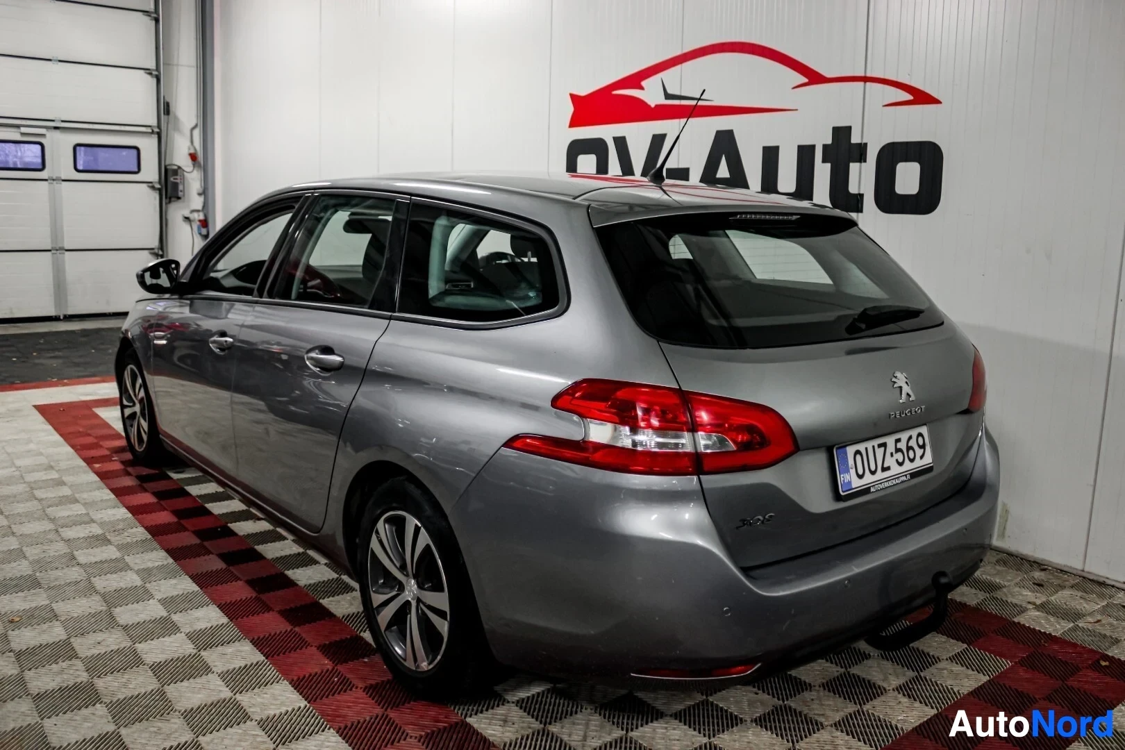 Takaviisto: Harmaa Peugeot 308 -farmariauto takaa ja oikealta, kuvattuna sisätiloissa ruudullisella lattialla ja OV-Auton logolla seinällä. Autossa on vetokoukku.