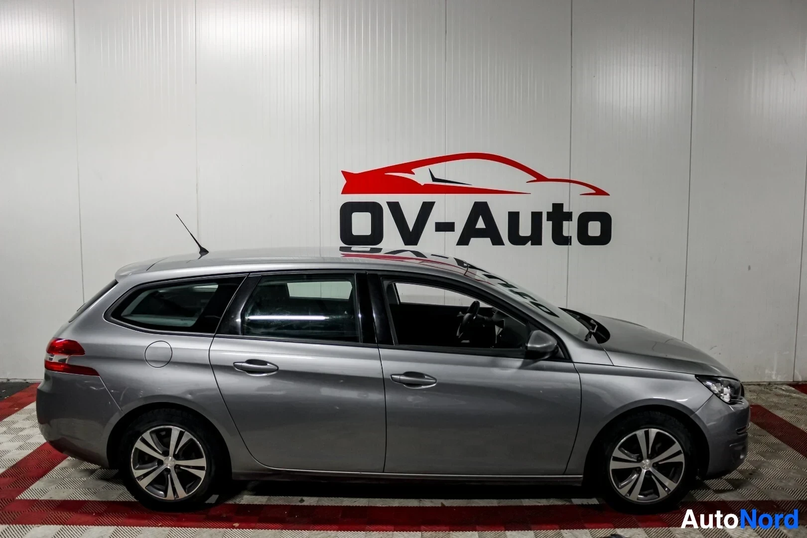Sivulta: Hopeanvärinen Peugeot 308 -farmariauto kuvattuna vasemmalta sivulta sisätiloissa. Autossa on kattokiskot ja monipuoliset kevytmetallivanteet. Auto on pysäköity