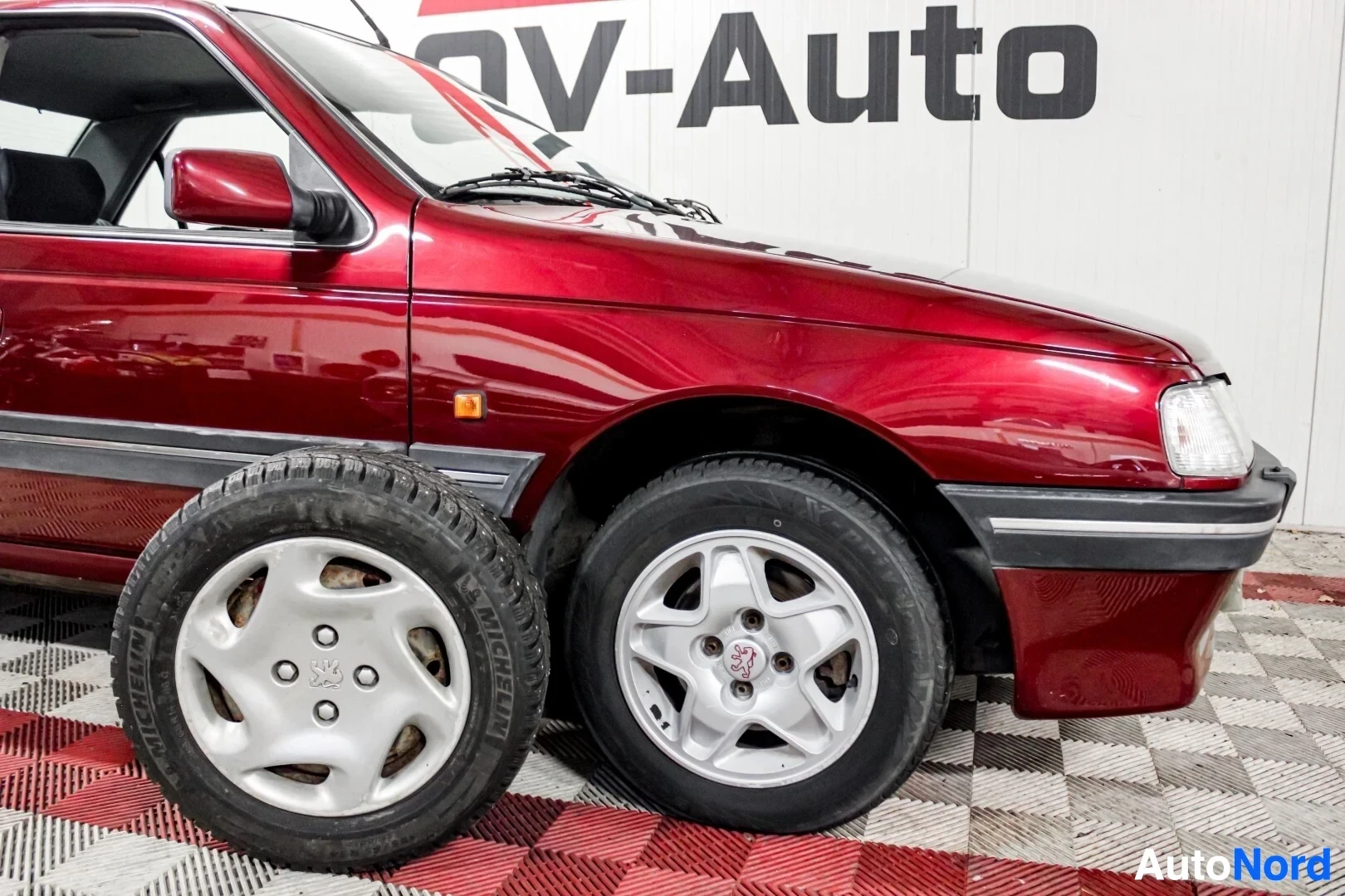 Etuviisto, nokka oikealle: Oikea etukulma tummanpunaisesta autosta, jossa on etupyörä ja toinen irtonainen pyörä, molemmat hopeisilla monipuolavanteilla. Auto seisoo ruutukuvioisel
