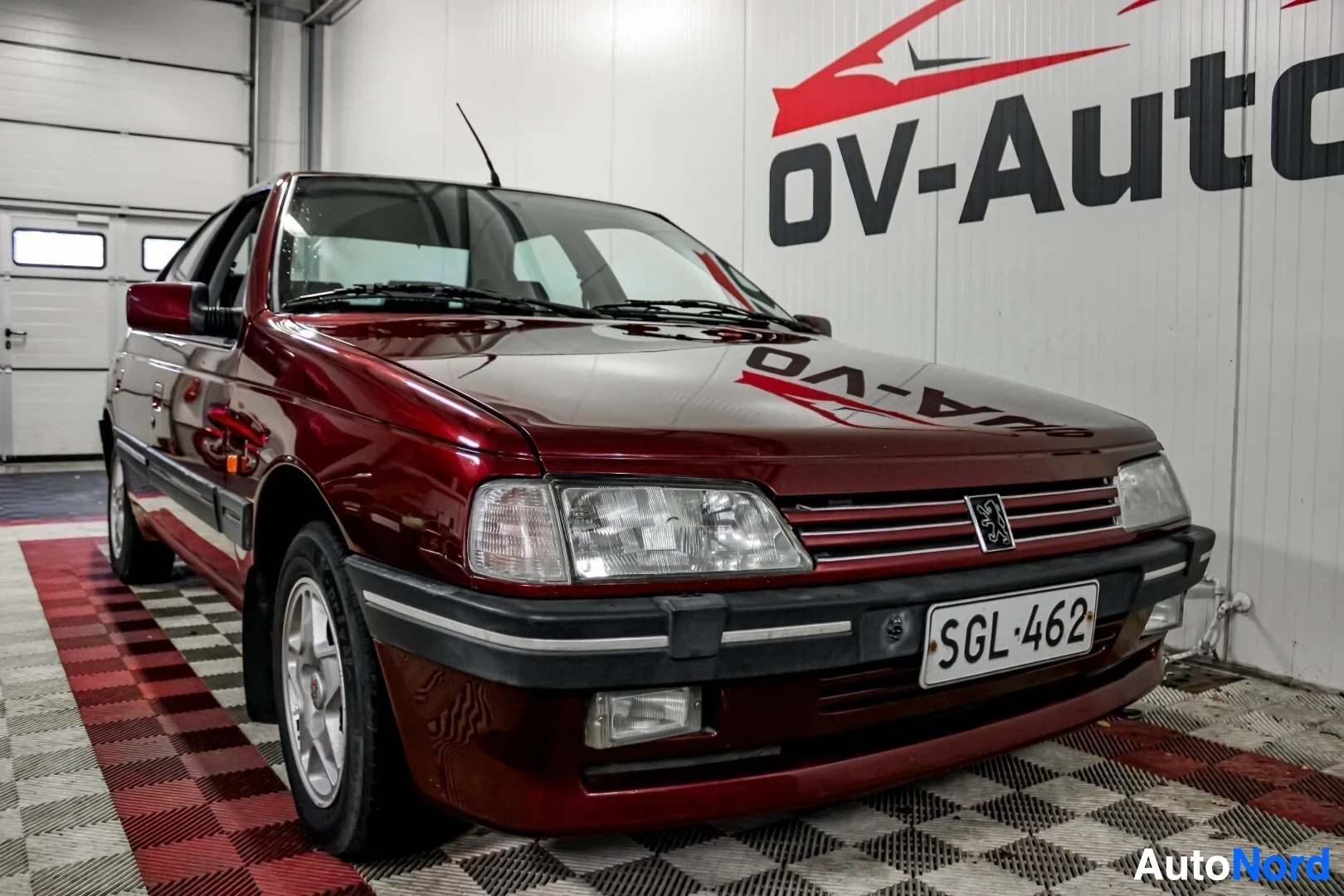 Etuviisto, nokka oikealle: Punainen Peugeot 405 edestä ja oikealta sivulta kuvattuna autokaupan sisätiloissa. Autossa on hopeanväriset vanteet ja rekisterikilpi