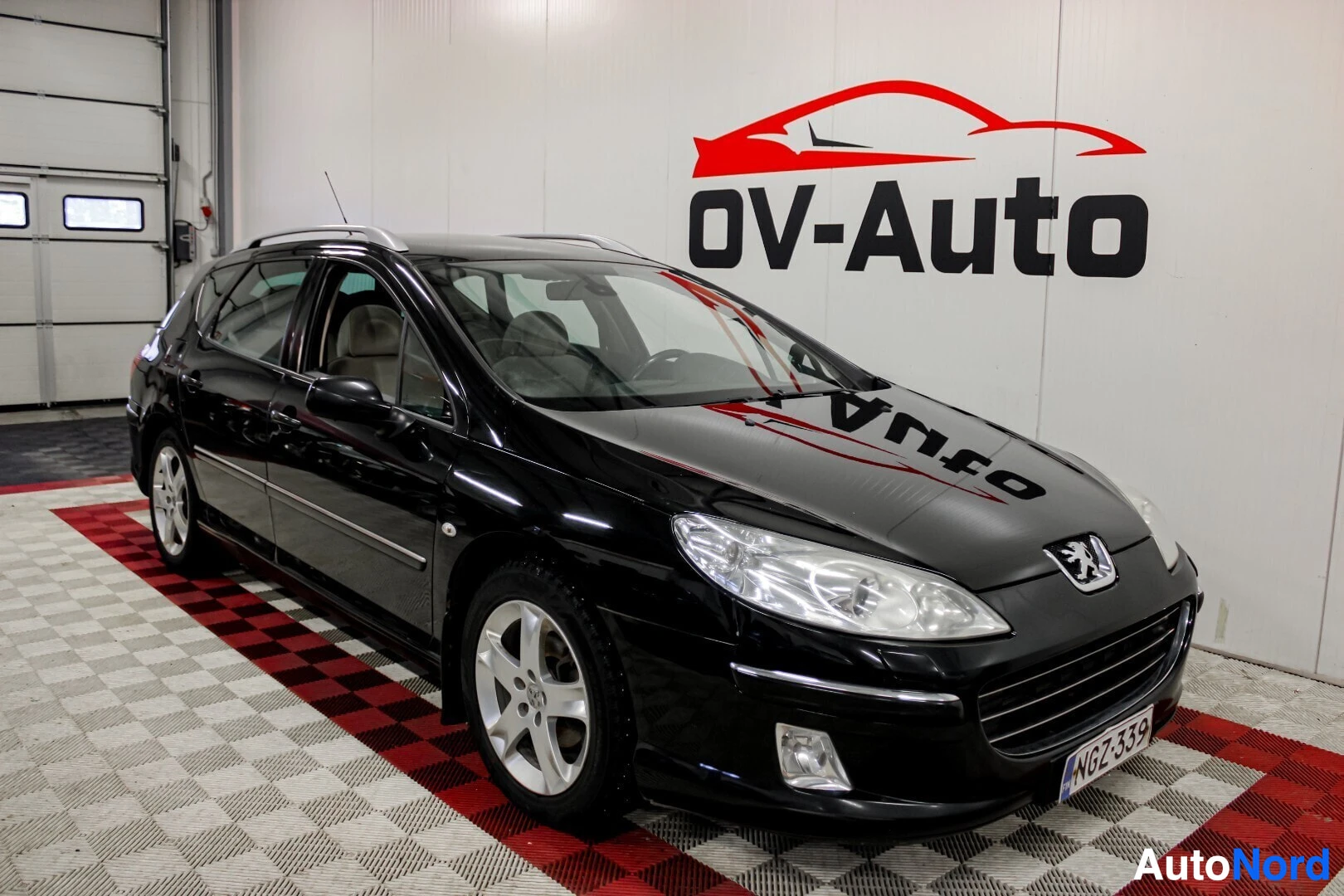 Etuviisto, nokka oikealle: Musta Peugeot 407 SW pysäköitynä valko-punaiseen ruutukuvioituun lattiaan, OV-Auto-logon edessä. Auton etu- ja oikea kylki näkyvät.