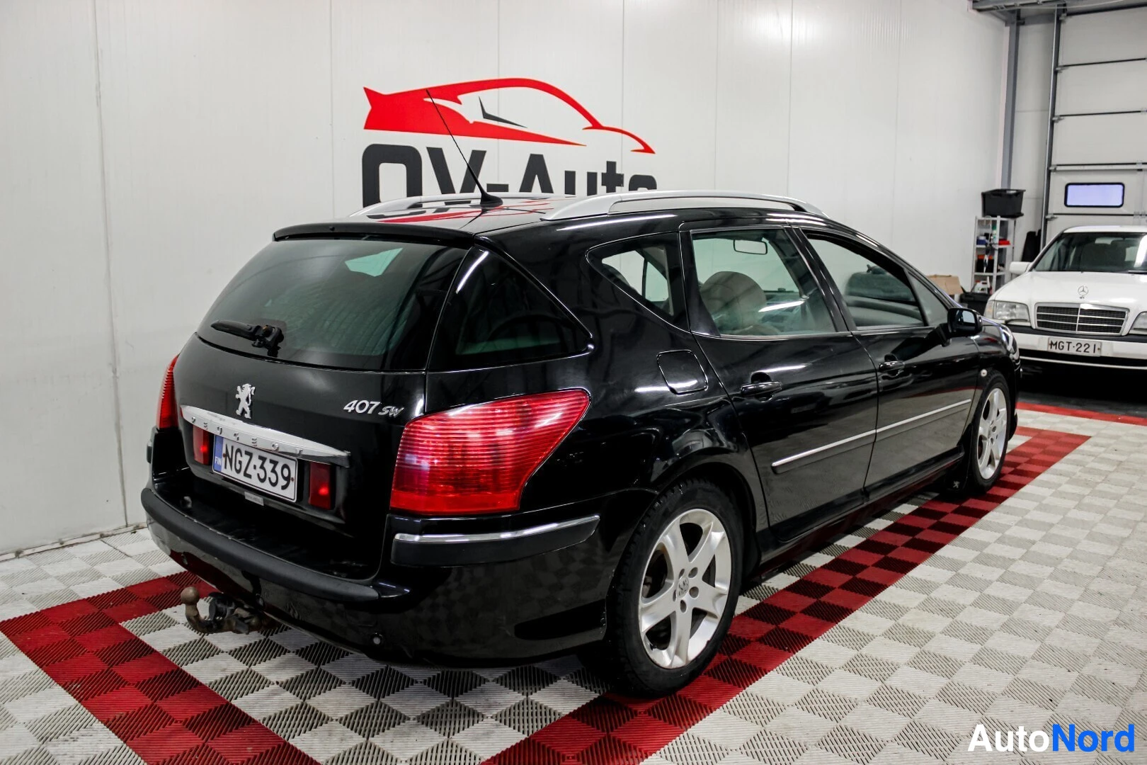 Takaviisto: Musta Peugeot 407 SW -farmariauto kuvattuna takaviistosta oikealta puolelta autohallissa. Autossa on kattokaiteet ja vetokoukku.