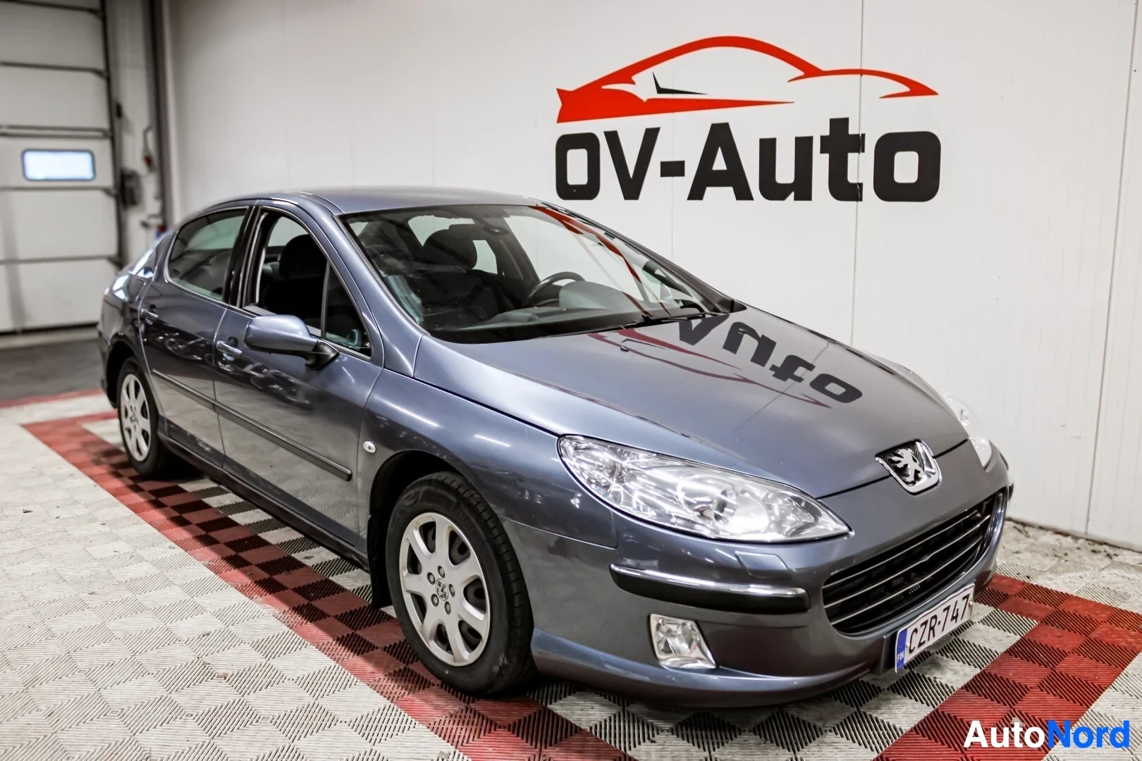 Etuviisto, nokka vasemmalle: Harmaa Peugeot 407 Sedan pysäköitynä sisätiloissa ruutukuvioidulla lattialla. Ajoneuvon etuosa ja vasen kylki ovat näkyvissä. Kuvassa näkyy osa rekiste