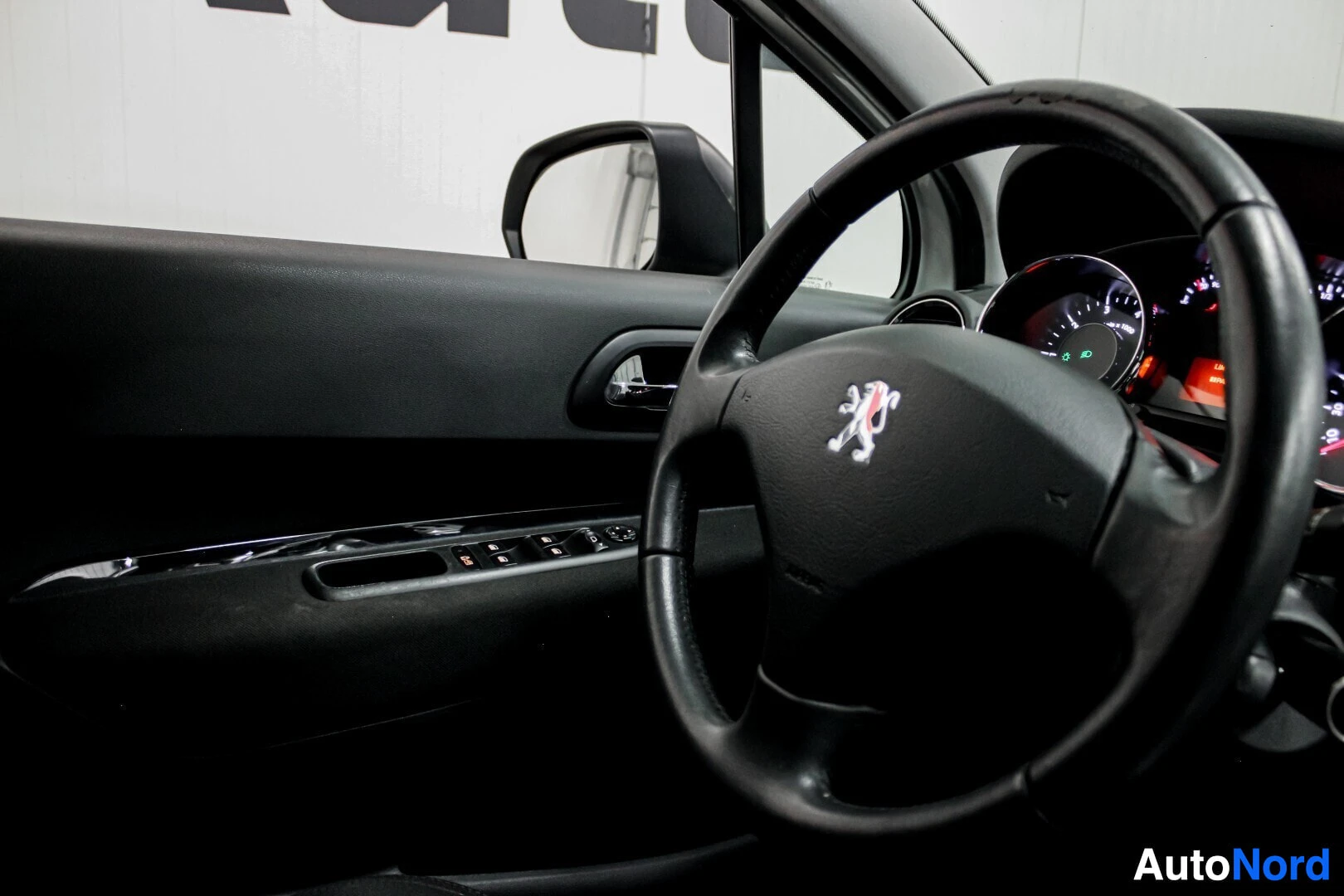 Ohjaamo: Ohjauspyörä, jossa on Peugeot-logo, ja musta ovipaneeli, jossa on ikkunoiden ja peilien säätimet. Ulkopeili näkyy osittain.