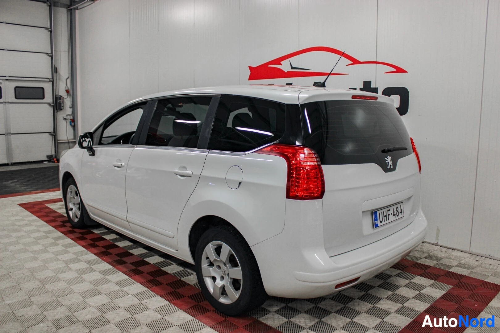 Takaviisto: Valkoinen Peugeot 5008 -henkilöauto pysäköitynä viistosti oikealle valkoiseen autohalliin, jossa on ruudullinen lattia. Auton takaosa ja oikea kylki näkyvät. Oi