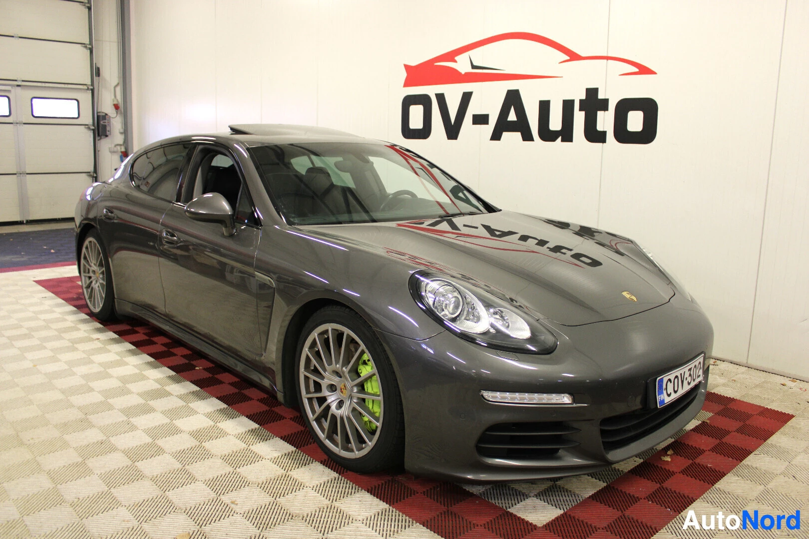 Etuviisto, nokka oikealle: Tummanharmaa Porsche Panamera S 3.0 V6 e-Hybrid edestä ja vasemmalta sivulta kuvattuna sisätiloissa. Autossa on kattoluukku ja hopeiset monipuolavanteet