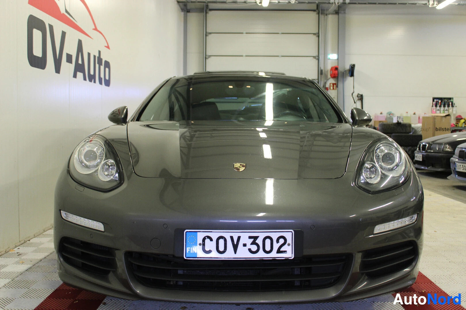 Edestä: Tummanharmaa Porsche Panamera S 3.0 V6 e-Hybrid edestä kuvattuna sisätiloissa. Autossa on kattoluukku. Rekisterikilpi on näkyvissä