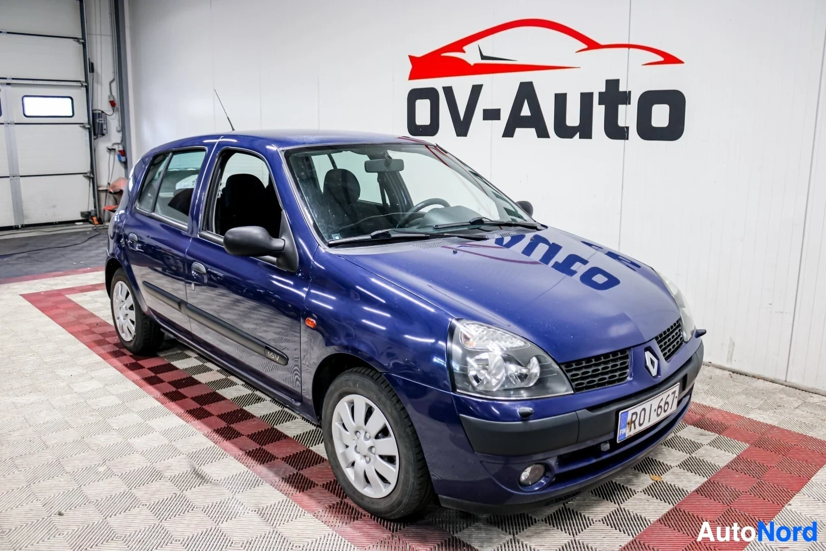 Etuviisto, nokka oikealle: Sinisellä metallivärillä maalattu Renault Clio on pysäköity halliin, jossa on OV-Auto-logo seinällä. Auton edessä ja oikealla puolella näkyy kuvassa. Aut