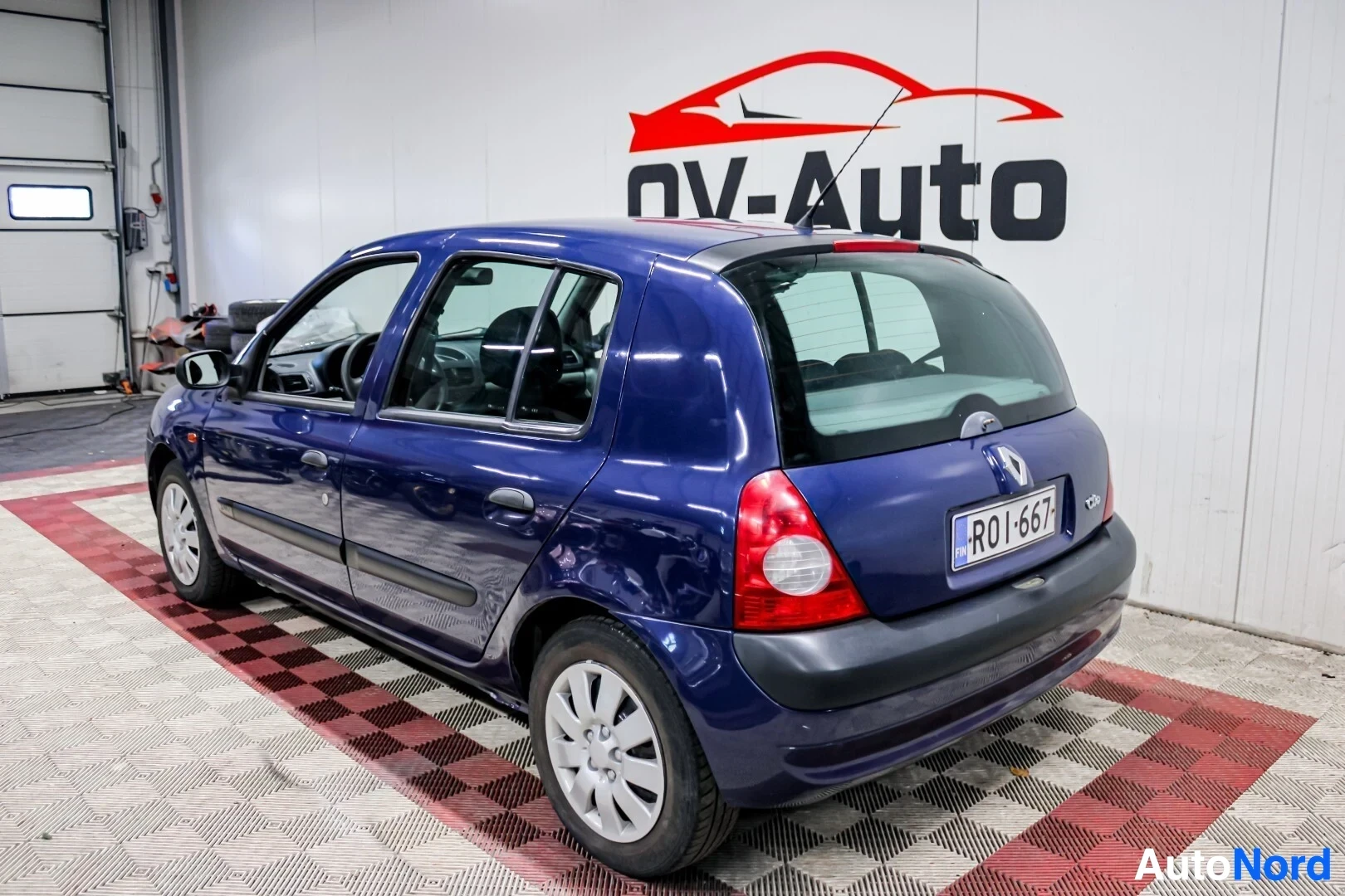 Takaviisto: Sinisellä metallivärillä maalattu Renault Clio on pysäköity halliin, jossa on OV-Auto-logo seinällä. Auton takana ja vasemmalla puolella näkyy kuvassa. Auton ta