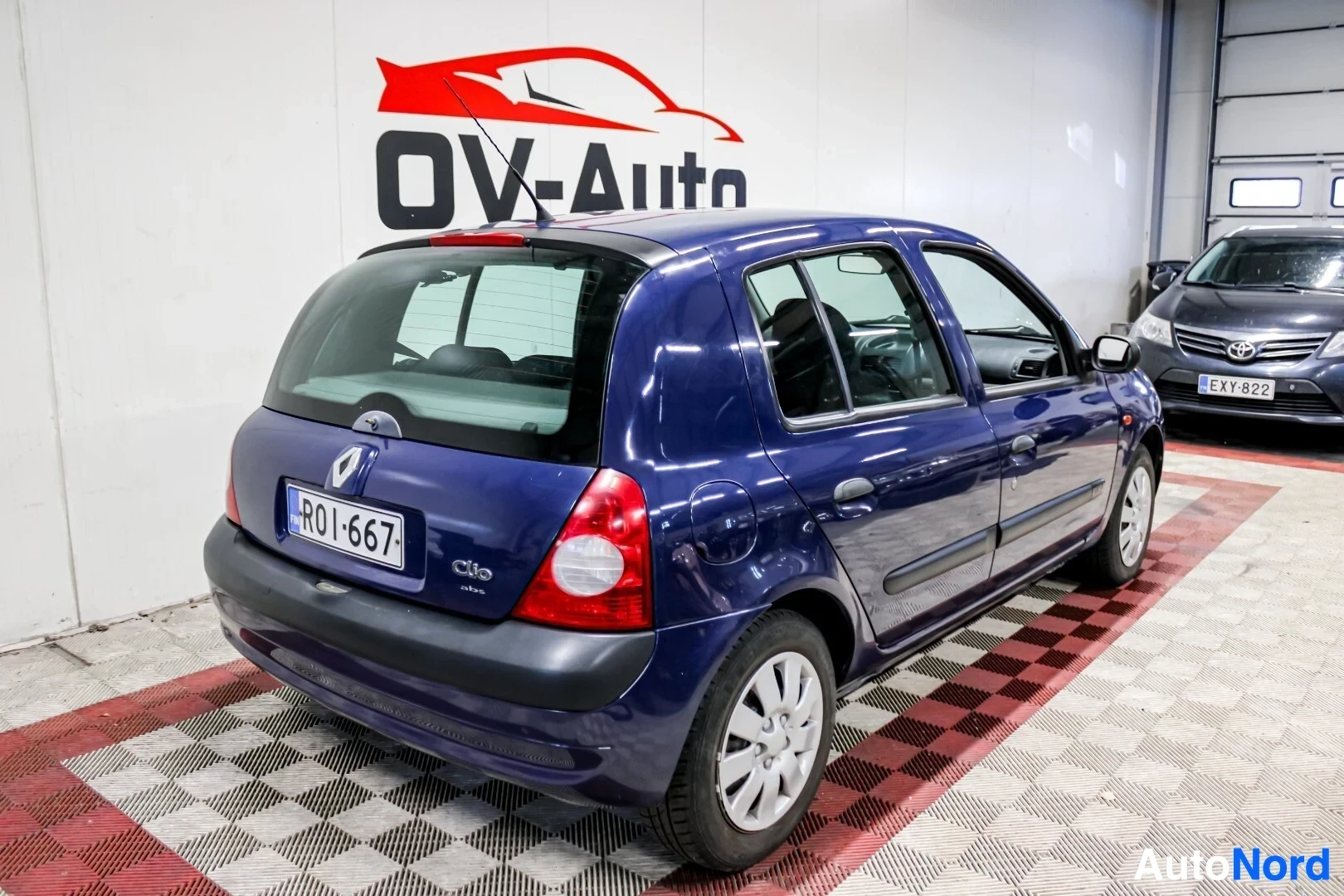 Takaviisto: Sininen viisiovinen hatchback-auto on pysäköity sisätiloihin. Auton takaosa ja vasen kylki ovat näkyvissä. Oikealla taustalla näkyy toinen auto, jonka rekisteri