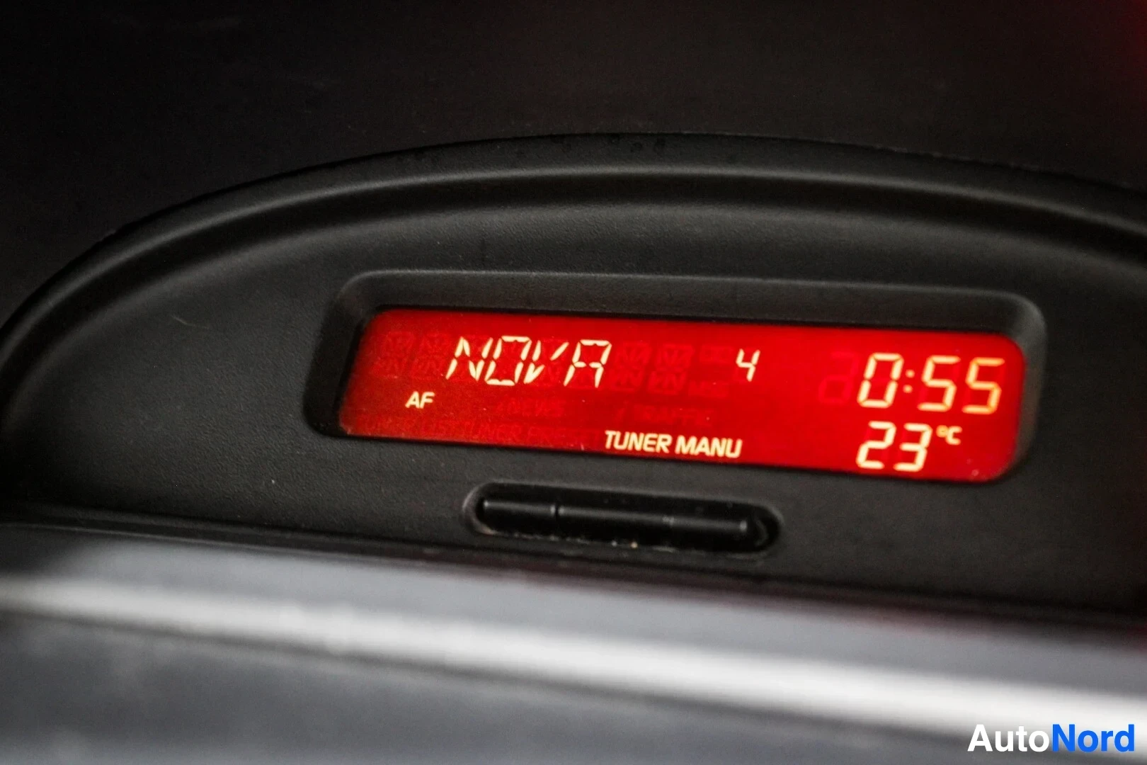 Yksityiskohta: Lähikuva auton digitaalisesta näytöstä, joka näyttää ajan "0:55", lämpötilan "23°C" ja tekstin "NOVA" ja "TUNER MANU".