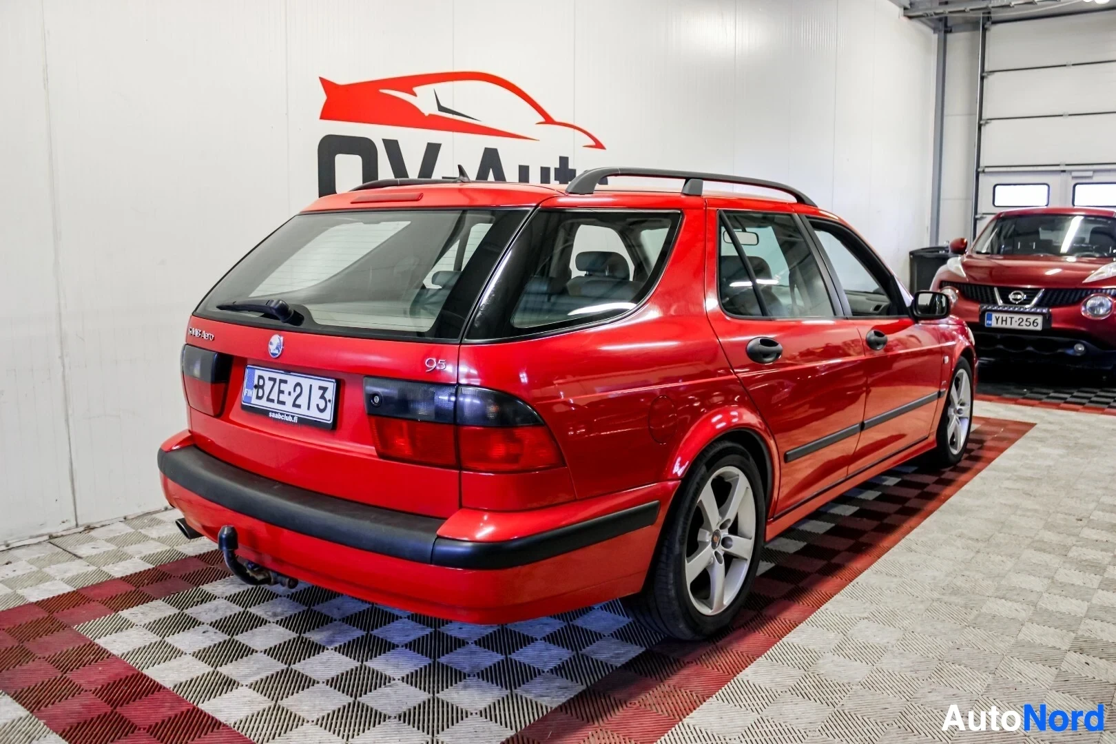 Takaviisto: Punainen Saab 9-5 farmari kuvattuna takaa oikealta kulmasta sisätiloissa. Autossa on kattokaiteet ja vetokoukku. Rekisterikilpi näkyy. Vieressä on toine