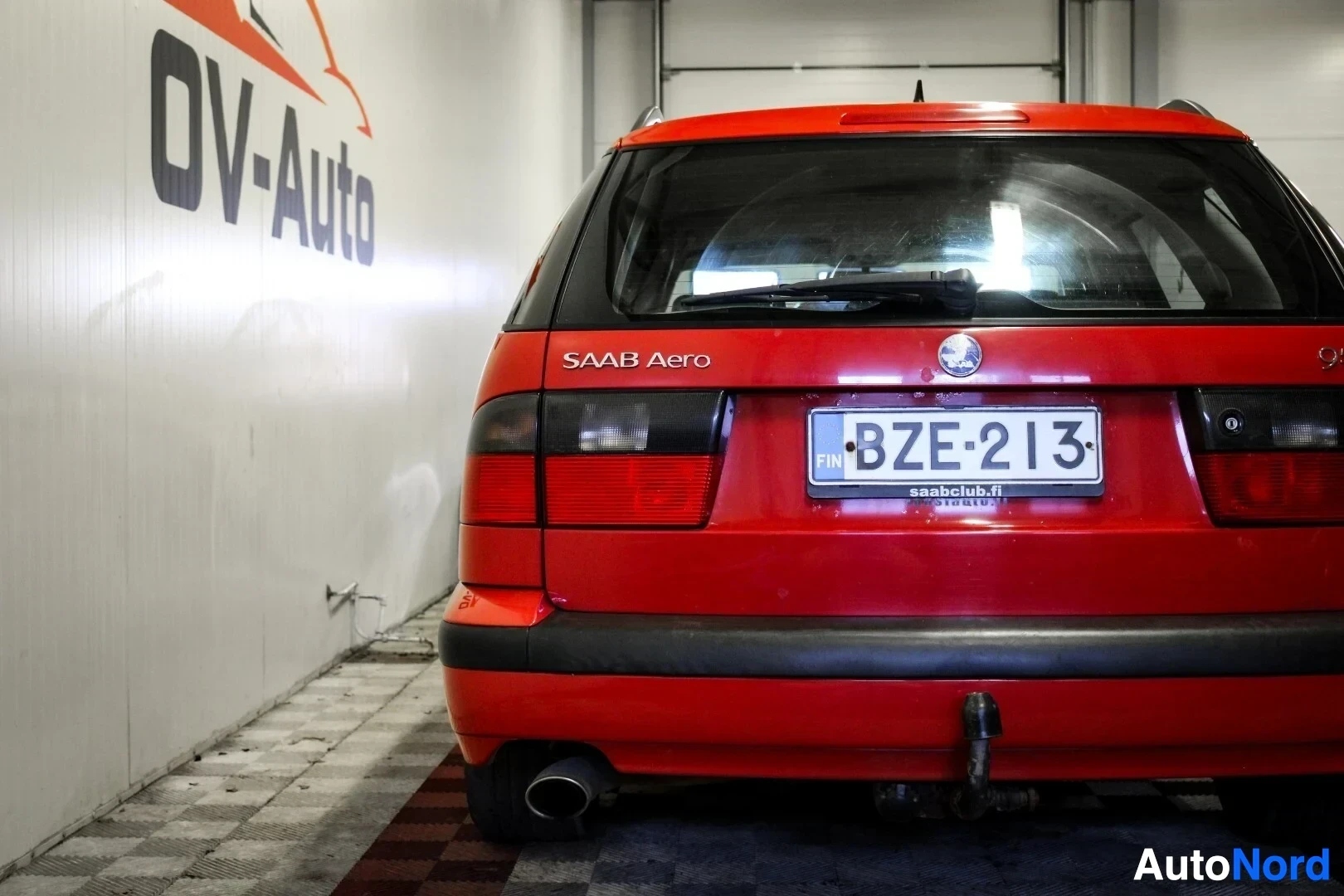 Takaa: Punaisen Saab 9-5 farmarin takaosa sisätiloissa. Vetokoukku ja rekisterikilpi näkyvät. Takalasissa on tarroja ja takaluukussa tekstit "SAAB Aero" ja "9