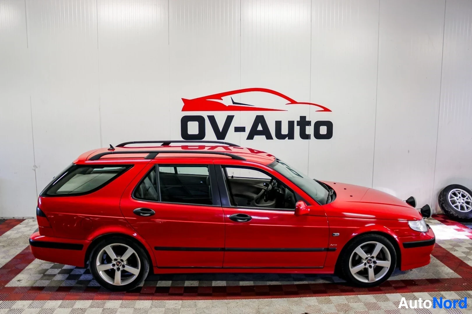 Sivulta: Punainen Saab 9-5 farmari kuvattuna oikealta sivulta sisätiloissa. Autossa on kattokaiteet ja kevytmetallivanteet. Taustalla näkyy OV-Auto-logo.