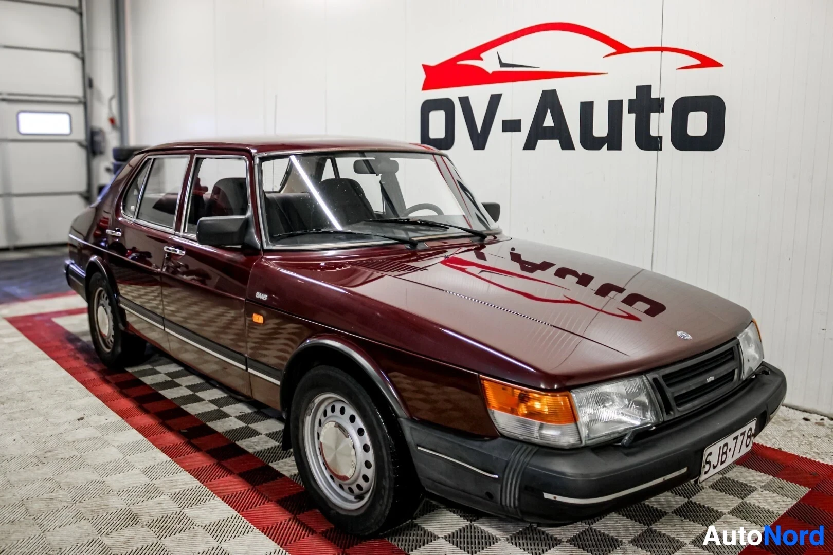 Etuviisto, nokka oikealle: Viininpunainen Saab 900 -henkilöauto, rekisteritunnus, pysäköitynä viistosti oikealle etuviistosta kuvattuna OV-Auto -kyltin edessä sisätiloissa