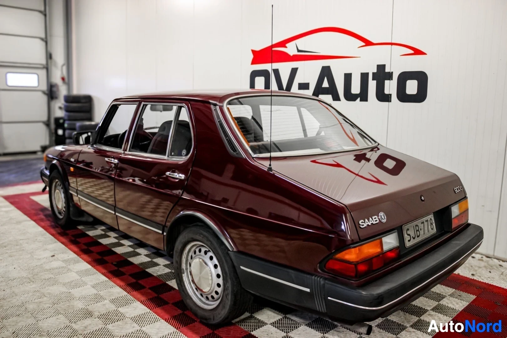 Takaviisto: Viininpunainen Saab 900 -henkilöauto, rekisteritunnus, pysäköitynä viistosti vasemmalle takaviistosta kuvattuna OV-Auto -kyltin edessä sisätiloissa
