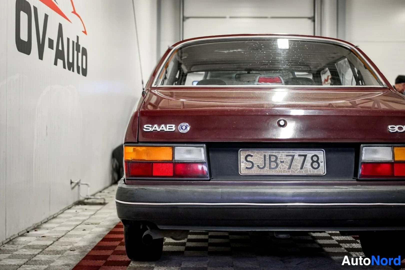 Takaa: Viininpunaisen henkilöauton takaosa, rekisterikilvessä teksti. Auto on kuvattu valkoisessa hallissa, jossa on OV-Auto-logo seinällä