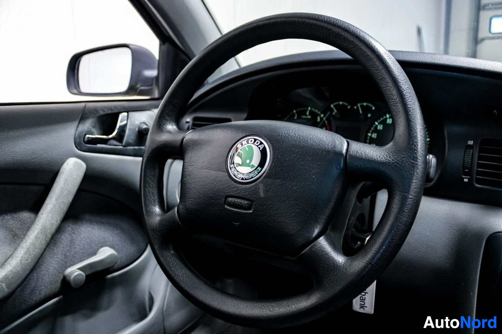 Ohjaamo: Kuvassa näkyy auton ohjauspyörä lähietäisyydeltä. Ohjauspyörä on musta, kolmipuolainen, ja sen keskellä on Skoda-logo sekä "AIRBAG"-teksti. Taustalla näkyy auto