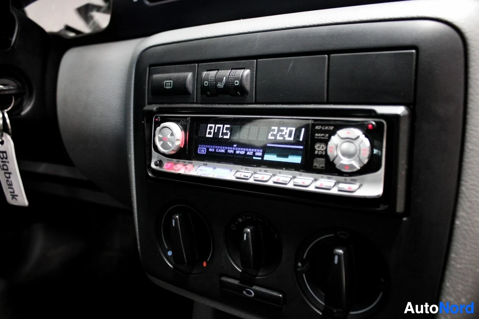 Yksityiskohta: Kuvassa on auton kojelaudan keskikonsoli. Siinä on jälkiasennettu autostereo, jossa näkyy numerot "87.5" ja "22:01". Stereon alapuolella on kolme pyöreää säädin