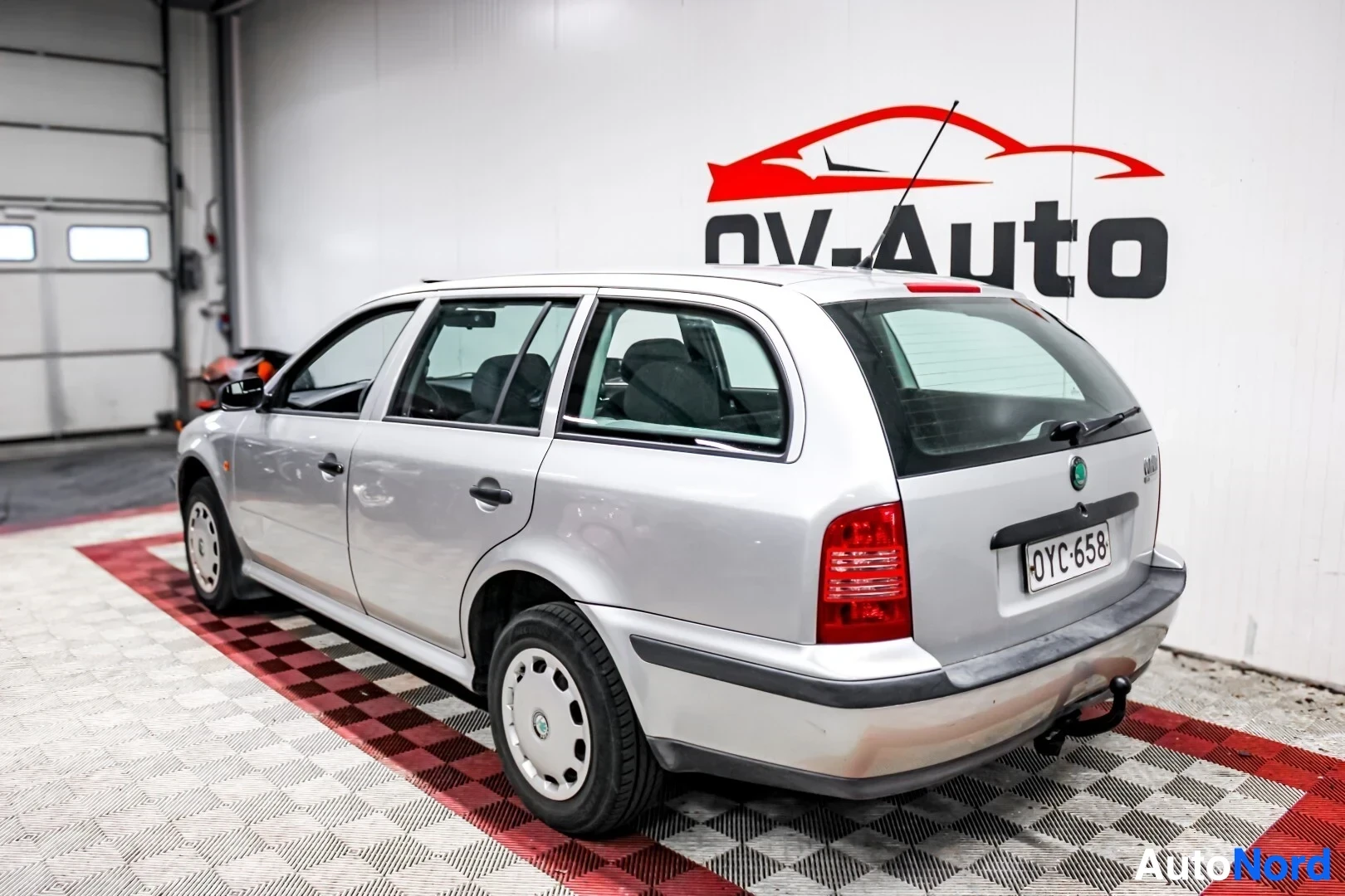 Takaviisto: Hopeanvärinen Skoda Octavia Combi -henkilöauto pysäköitynä viistosti oikealta takaa kuvattuna. Autossa on kattokaiteet ja vetokoukku. Rekisterikilpi on