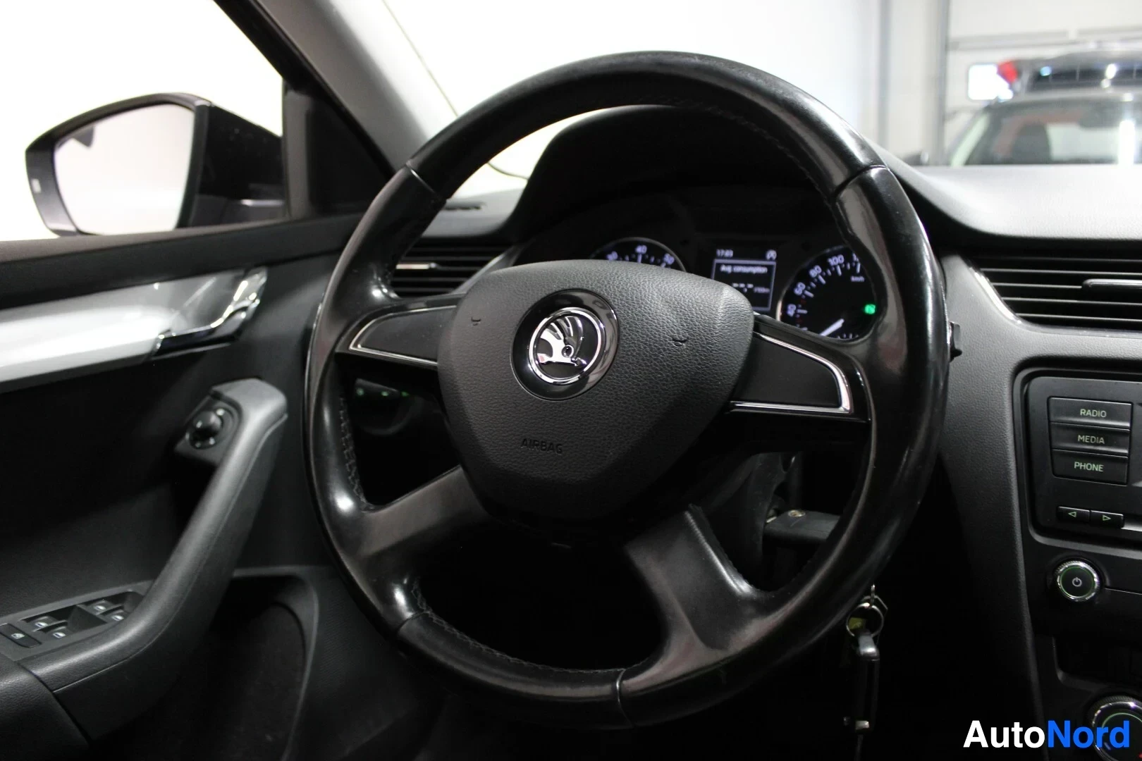 Ohjaamo: Lähikuva ohjauspyörästä, jossa on Skoda-logo ja AIRBAG-merkintä, sekä auton radiosta ja kojelaudasta taustalla.