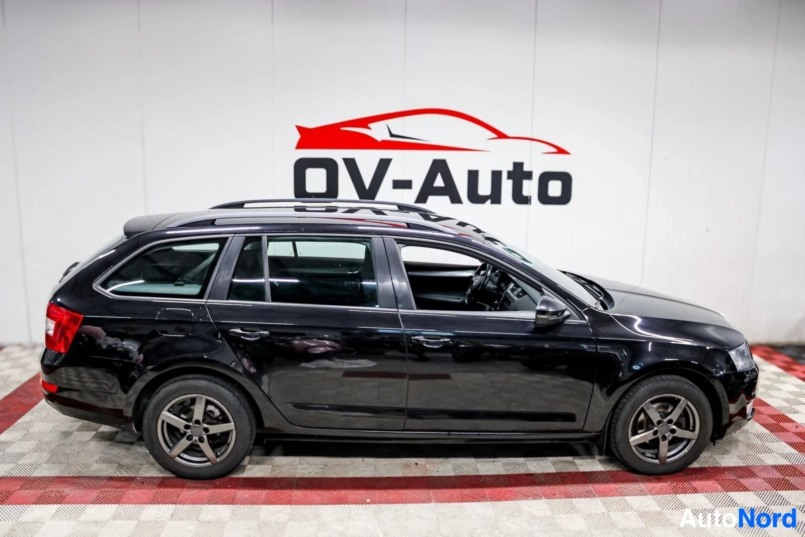 Sivulta: Musta Skoda Octavia farmari auto kuvattuna vasemmalta sivulta. Autossa on kattokiskot ja tummanharmaat kevytmetallivanteet.