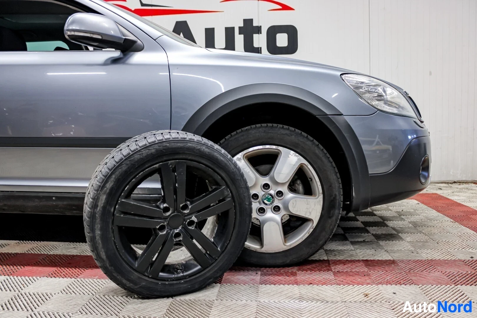 Etuviisto, nokka vasemmalle: Auton etuosa oikealta puolelta, etuvalo, pyörä ja rengas sekä toinen musta vanne ja rengas edessä. Auton kyljessä lukee "AUTO".