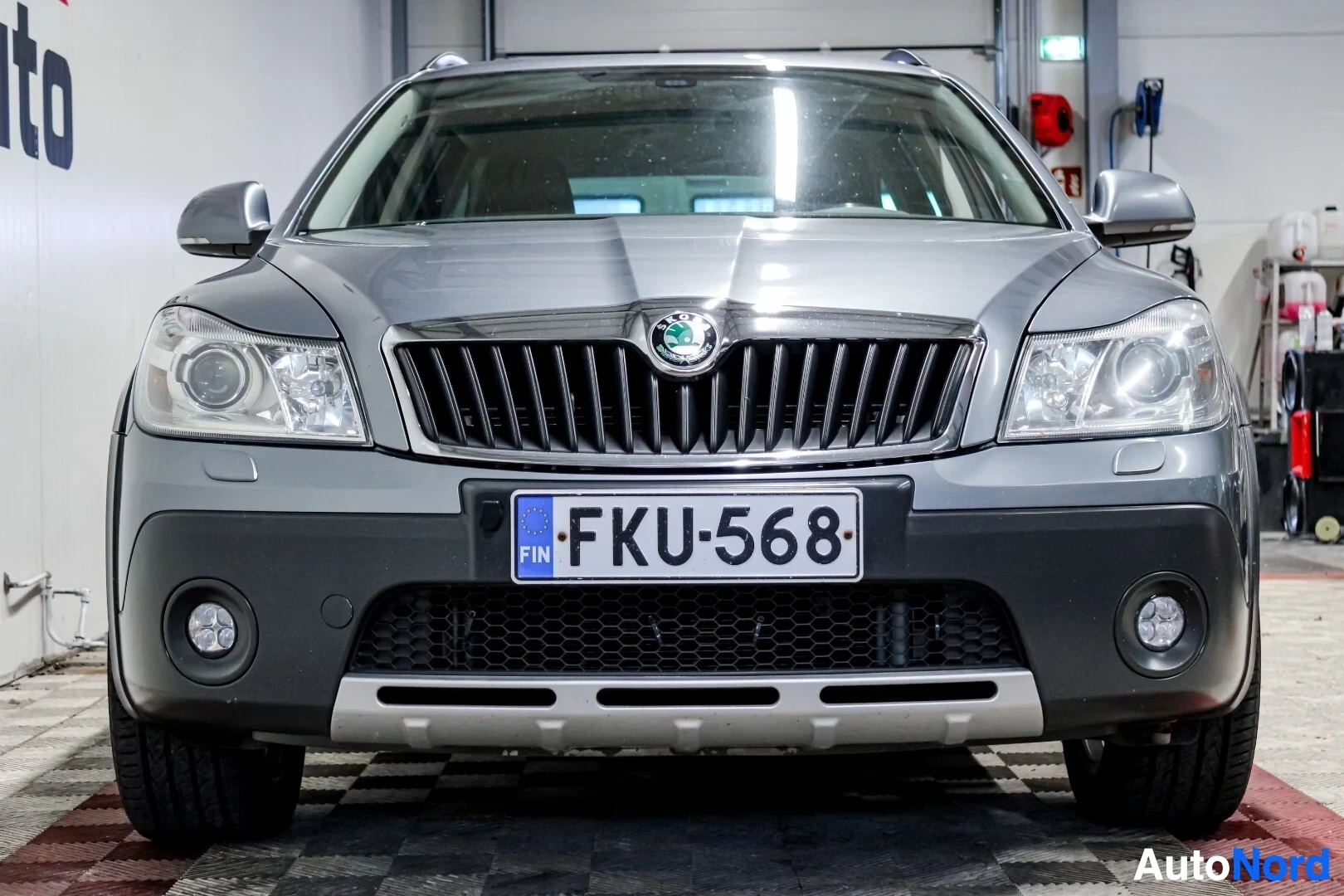 Edestä: Harmaa farmariauto edestä päin kuvattuna autohallissa. Ajovalot, etusäleikkö, Skodan logo ja rekisterikilpi "" ovat keskellä kuvaa. Etupuskurissa on kaks