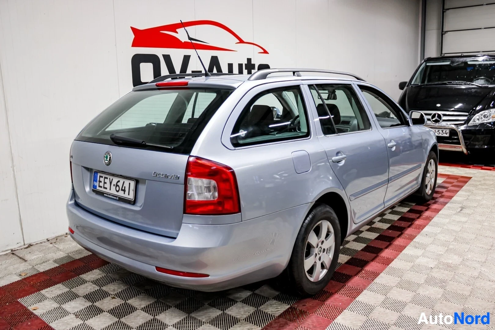 Takaviisto: Vaaleansininen farmariauto takaviistosta oikealta kuvattuna, jossa näkyy auton perä ja oikea kylki. Autossa on kattokiskot ja takaluukussa kilpi