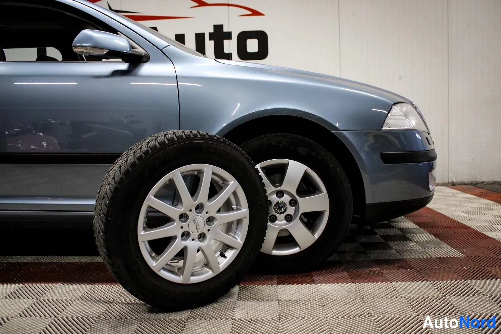 Etuviisto, nokka oikealle: Sinertävänharmaa Skoda Octavia kolmannelta kylkikulmalta, etuosa ja kylki näkyvissä; eturenkaan vieressä pöydällä vaihtopyörä hopeavanteella.