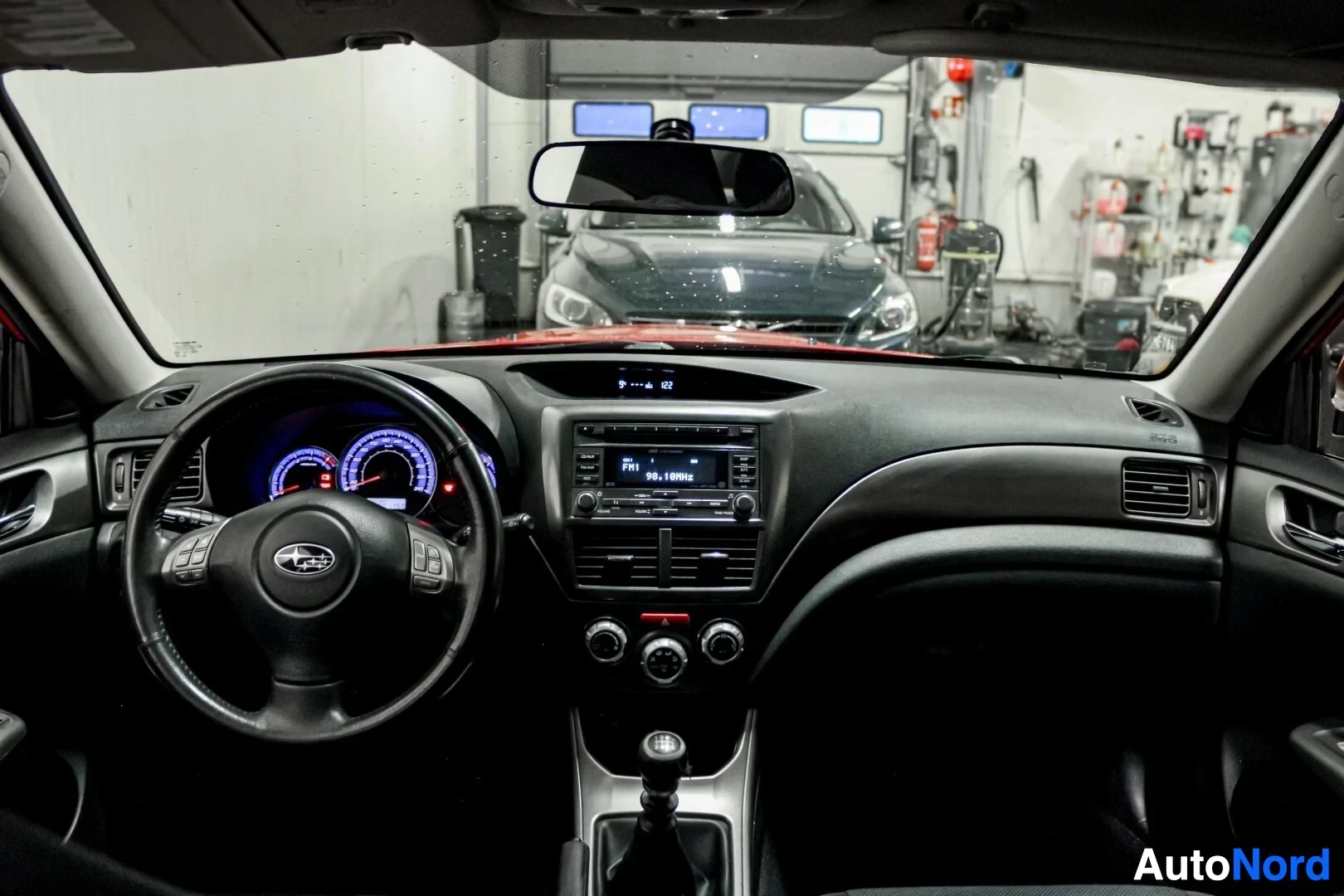 Ohjaamo: Kojelauta, ohjauspyörä ja mittaristo kuvattuna auton sisältä tuulilasin läpi. Taustalla näkyy toinen auto autohallissa.