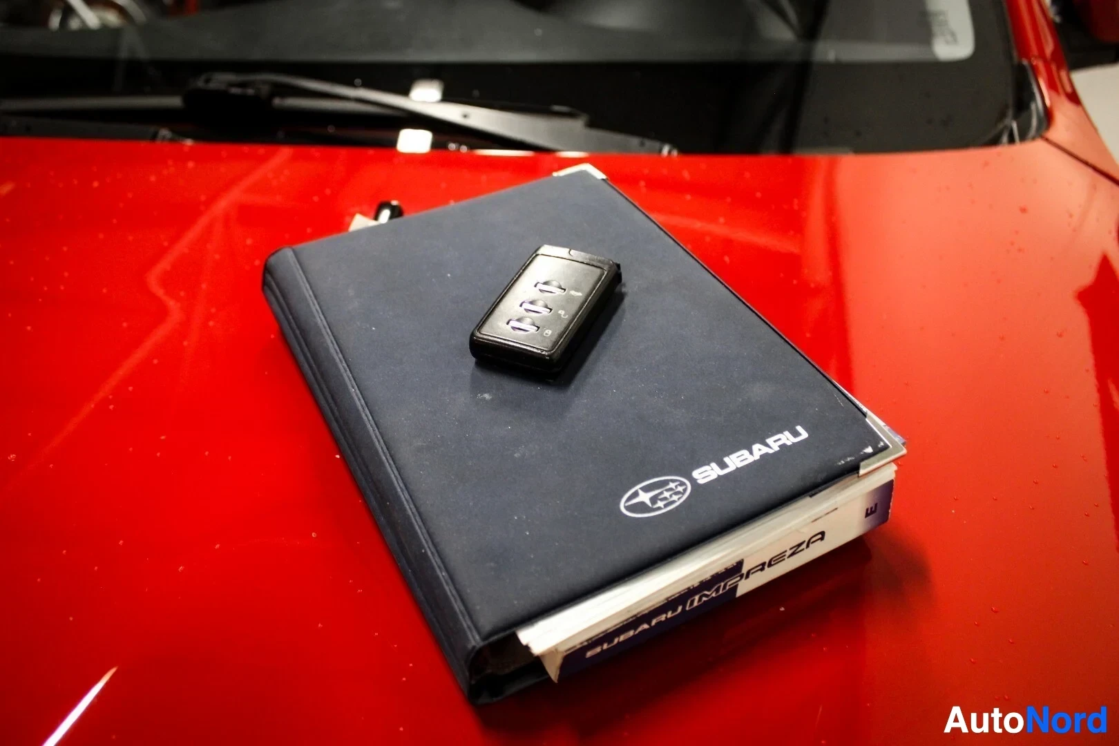 Yksityiskohta: Musta avaimenperä, jossa on painikkeita, lepää tummansinisen "SUBARU"-logolla varustetun kirjan päällä, joka on puolestaan punaisen auton konepellillä.