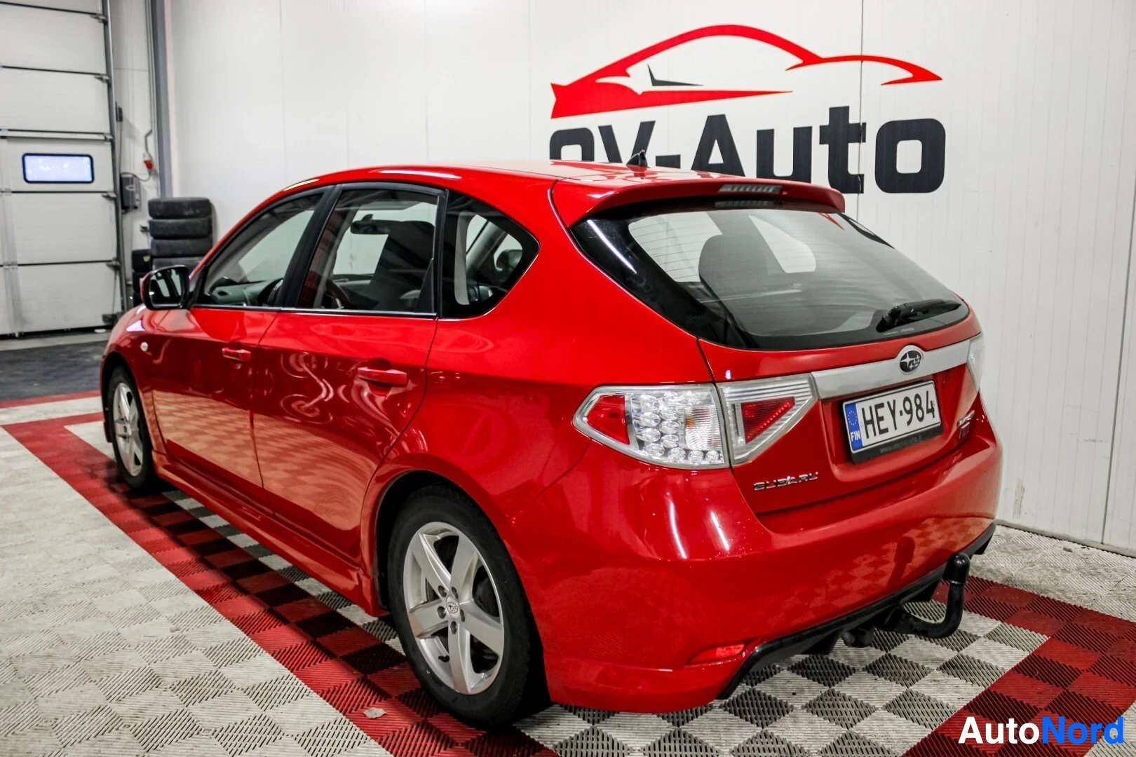 Takaviisto: Punainen viistoperäauto takaa ja sivulta päin kuvattuna sisätiloissa. Autossa on vetokoukku ja hopeiset kevytmetallivanteet. Taustalla näkyy OV-Auto-logo ja val