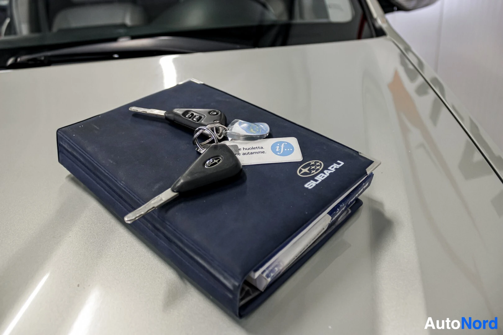 Yksityiskohta: Lähikuva auton konepellillä olevasta tummansinisestä Subaru-merkkisestä käsikirjasta ja kahdesta avaimesta, joissa toisessa on Subaru-logo.