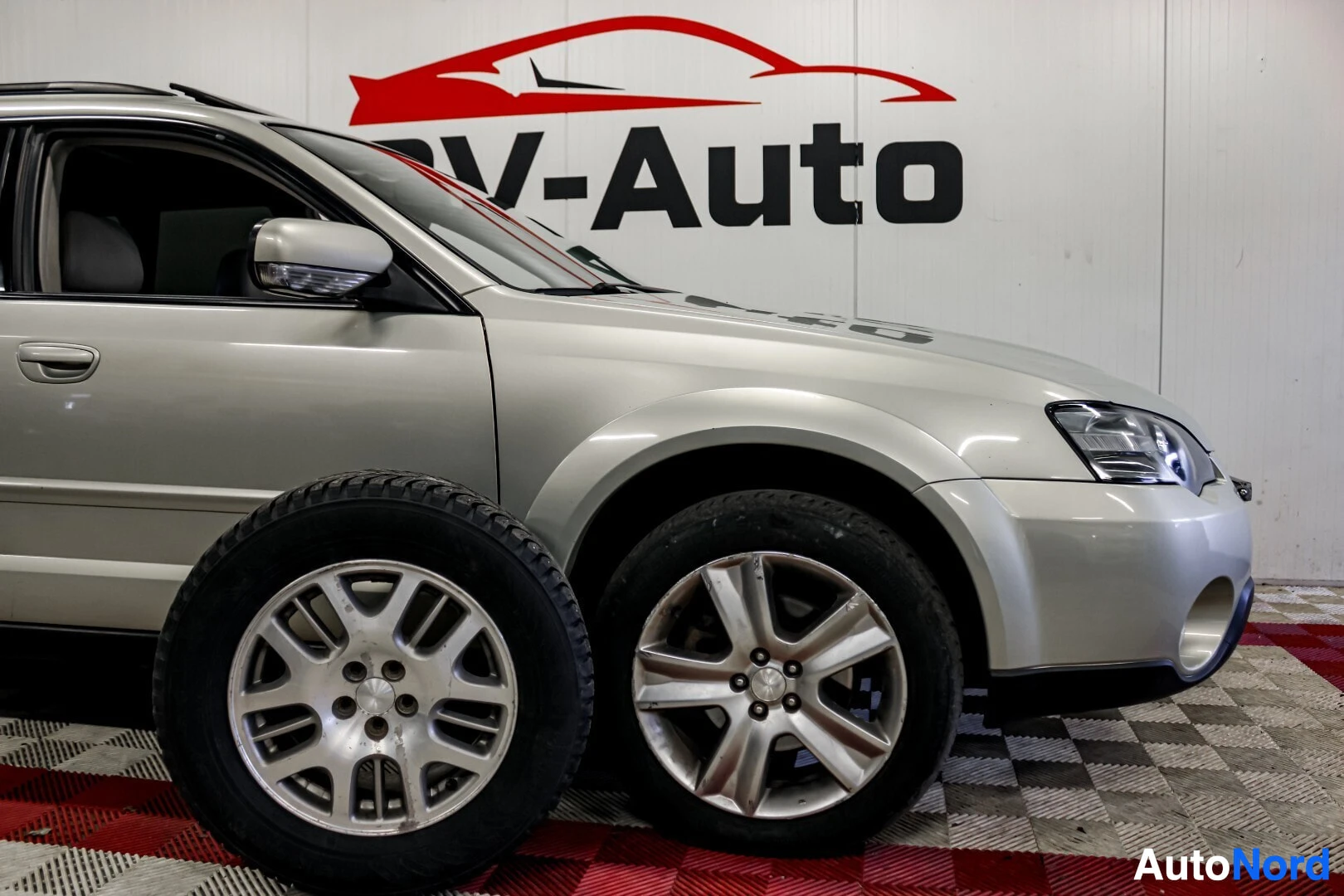 Etuviisto, nokka oikealle: Vaalea Subaru Outback seisoo etuvasemmalla kuvattuna. Sen vieressä on toinen rengas vanteineen. Auton takana näkyy seinä, jossa lukee "V-Auto".