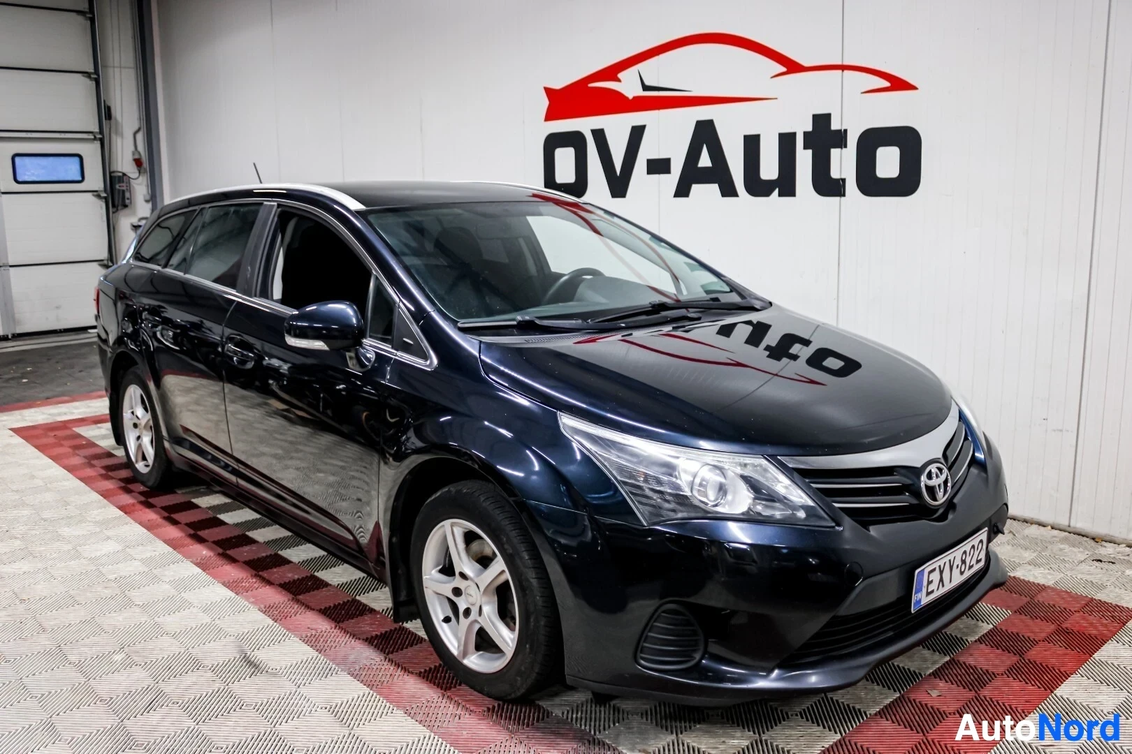 Etuviisto, nokka vasemmalle: Tumman yönsininen Toyota Avensis farmari kuvattuna etuviistosta oikealta sisätilassa. Autossa on kattokaiteet ja alumiinivanteet.