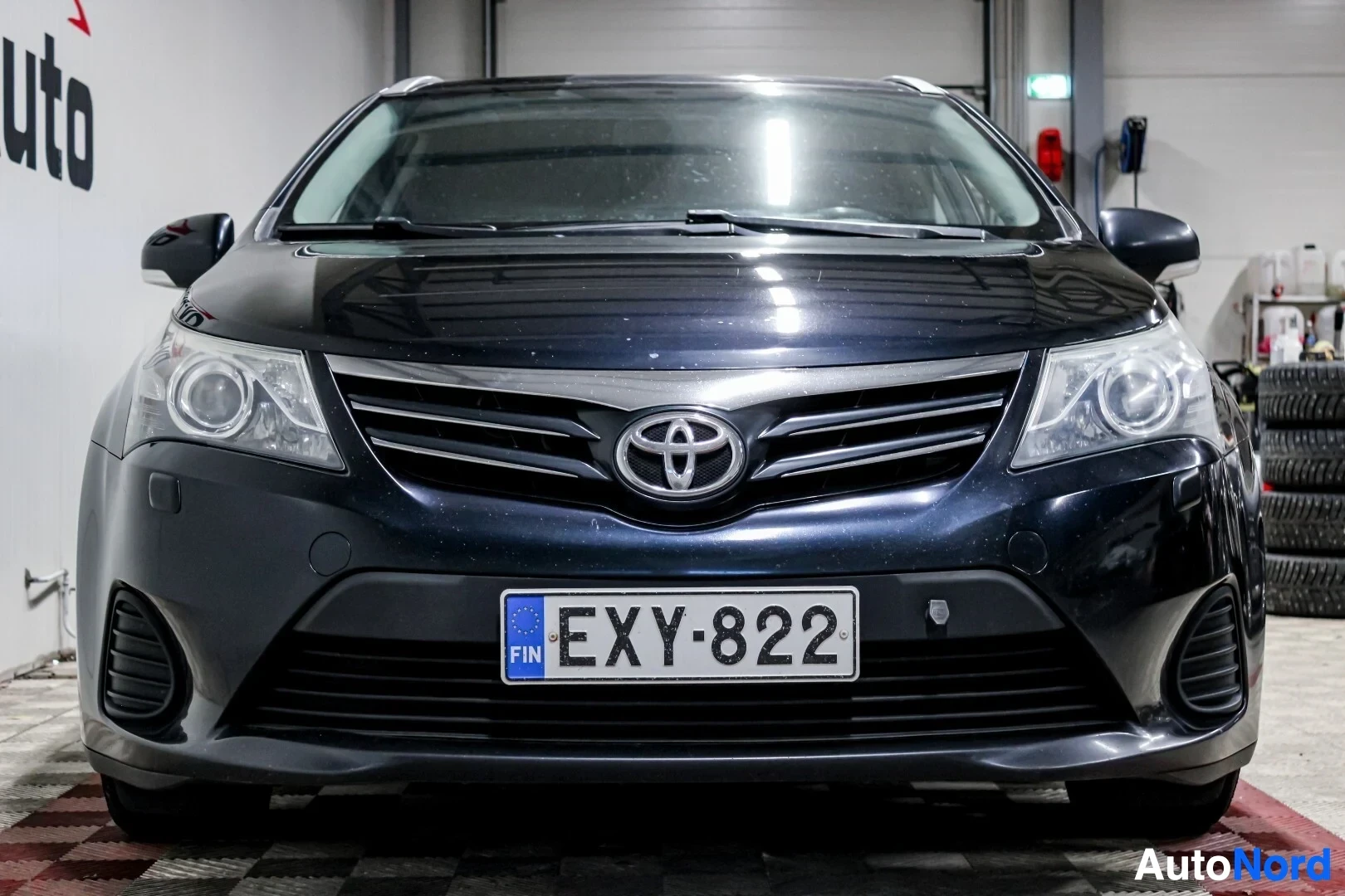 Edestä: Tumman yönsinisen Toyota Avensis farmariauton etuosa kuvattuna suoraan edestä sisätilassa. Rekisterikilpi on näkyvissä.