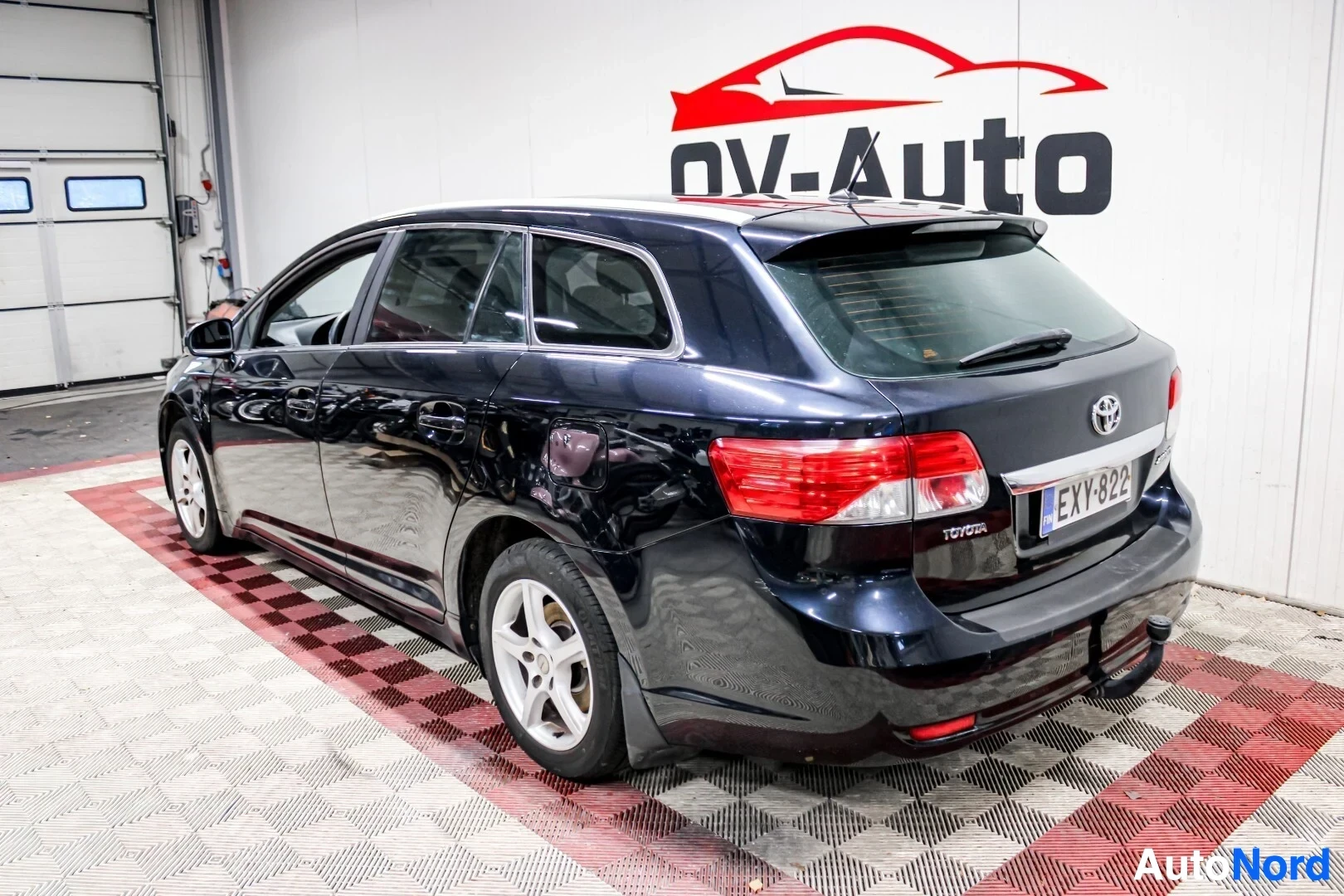 Takaviisto: Tumman yönsininen Toyota Avensis farmariauto kuvattuna takaviistosta vasemmalta sisätilassa. Autossa on vetokoukku, kattokaiteet ja alumiinivanteet.