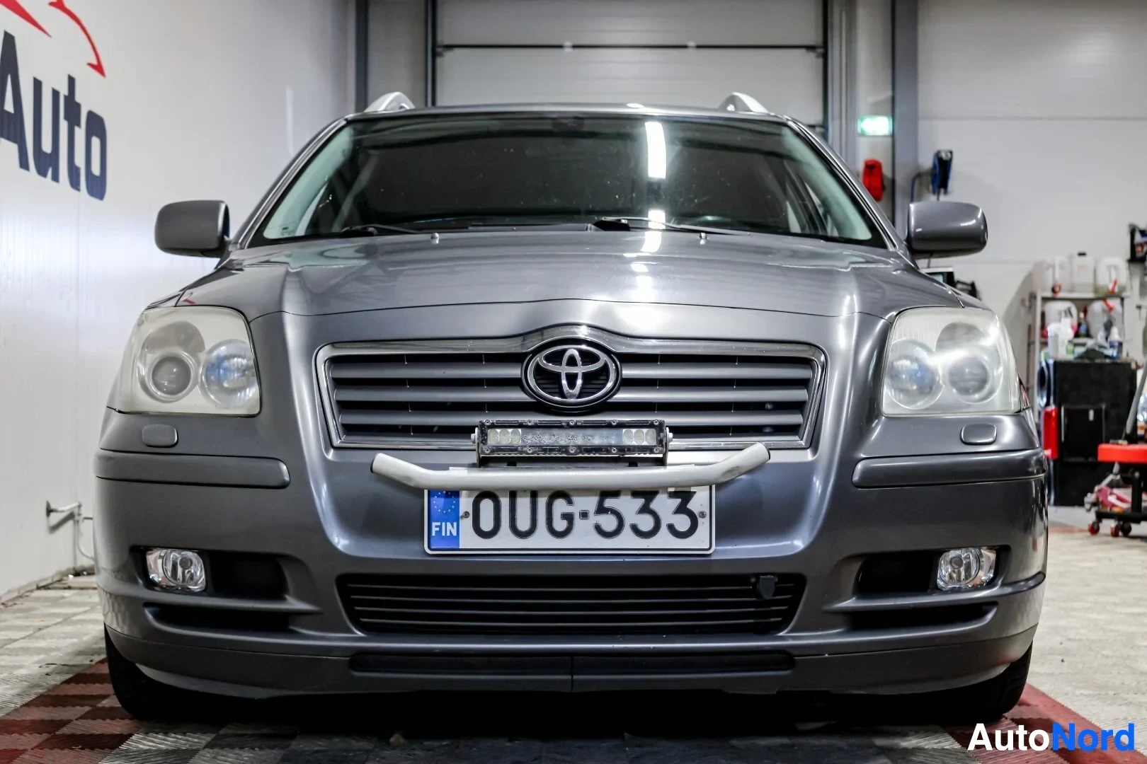 Edestä: Harmaa farmariauto edestä. Auton etuosassa on rekisterikilpi "" ja lisävalo kilven yläpuolella. Etusäleikössä on Toyota-logo