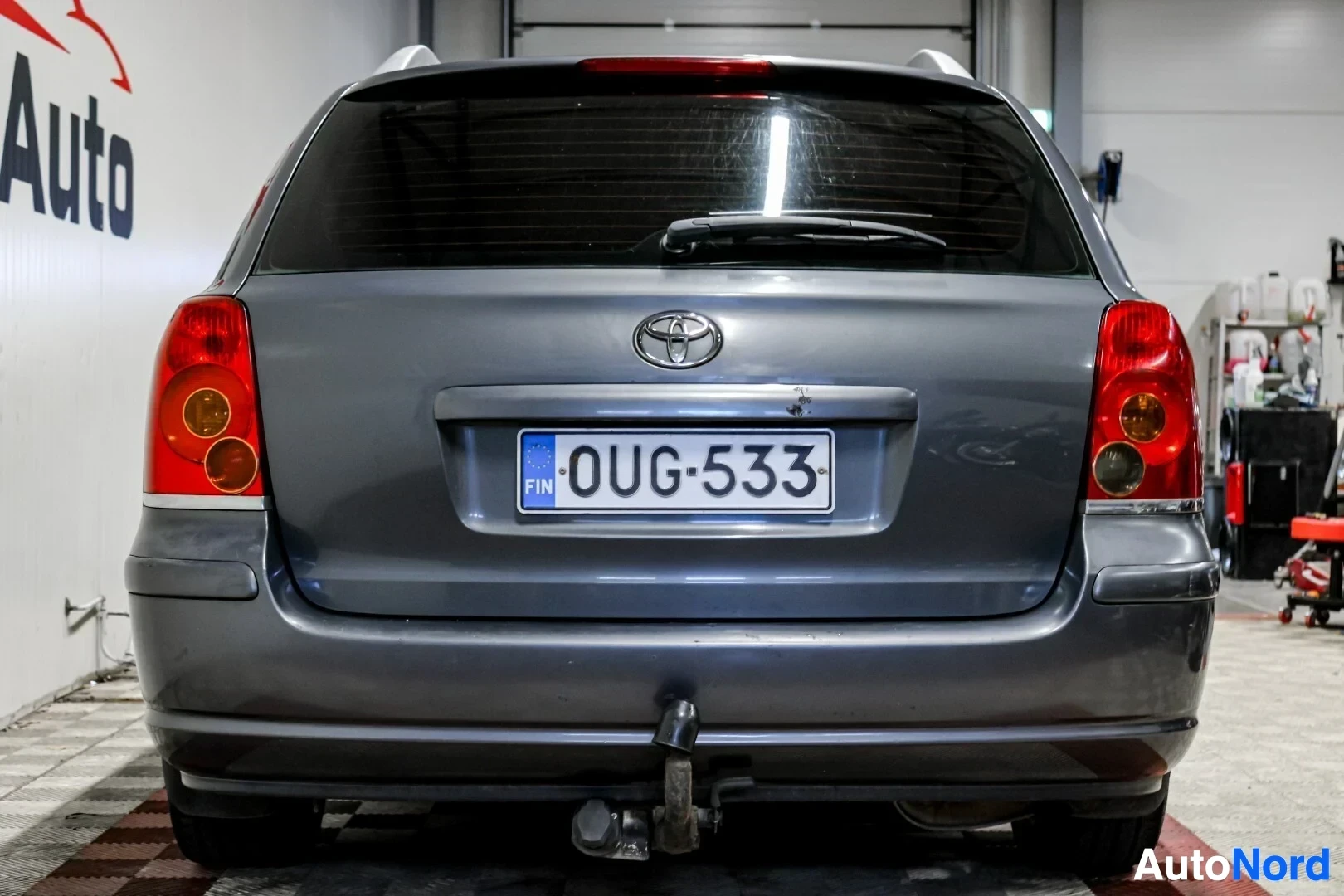 Takaa: Harmaa farmariauto, jossa on vetokoukku, pysäköitynä valkoisen seinän edessä. Auto on kuvattu suoraan takaa.