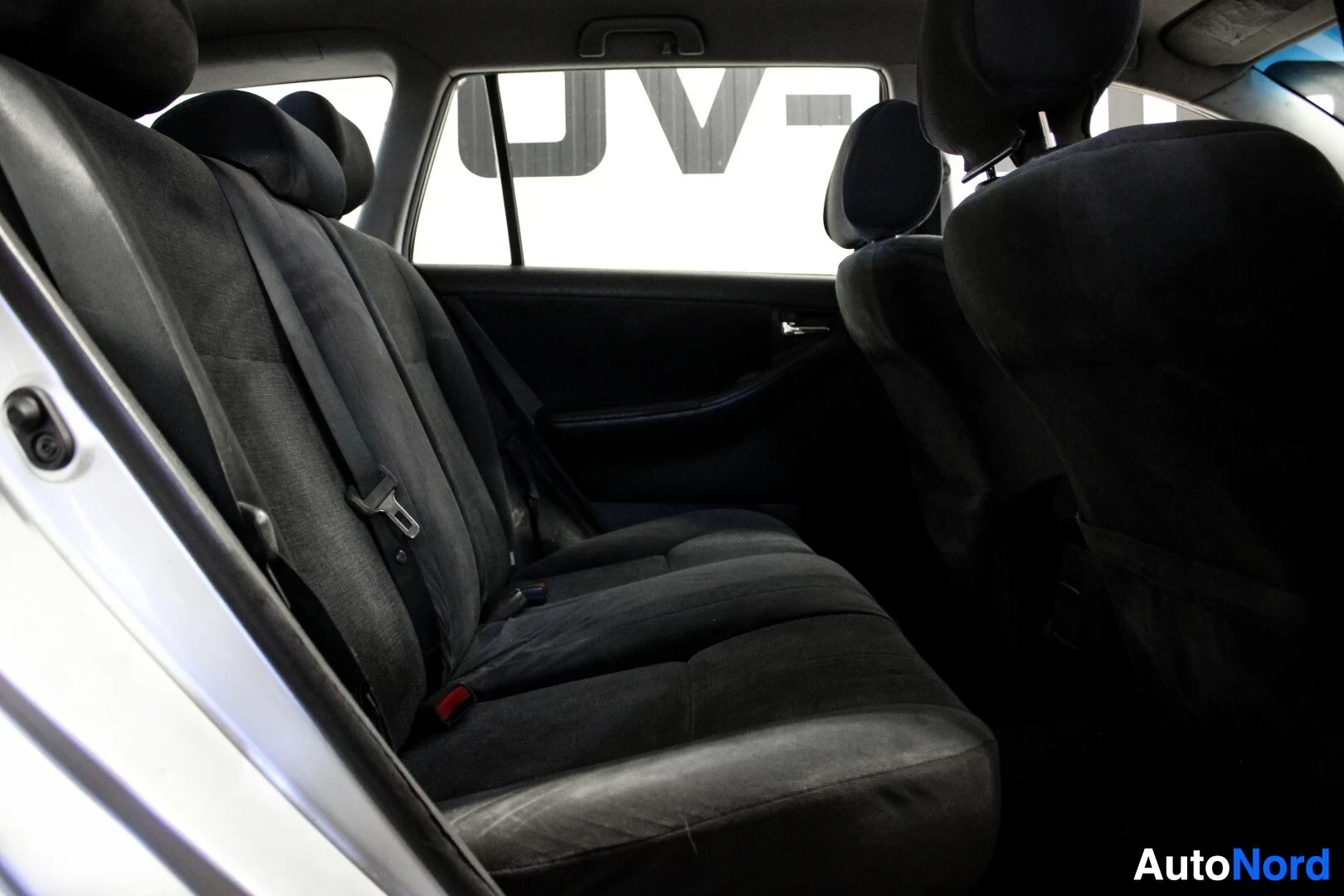 Takaistuimet: Takaikkunan läpi näkyy valkoinen autohallin seinä, ja takana on kangasverhoiltu istuin.
