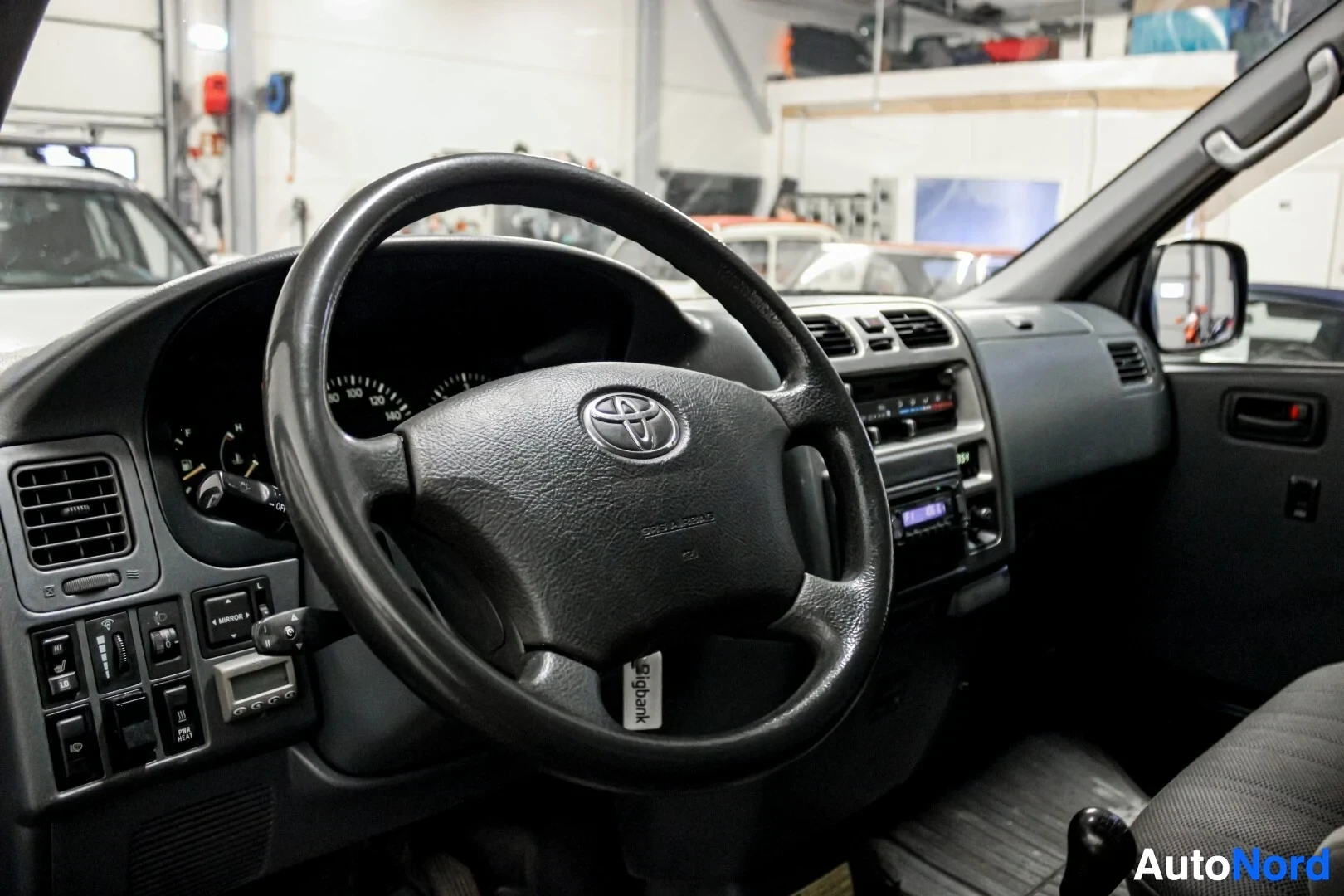 Yksityiskohta: Lähikuva ohjauspyörästä, jossa on Toyota-logo keskellä. Ohjauspyörä on musta ja kolmipuolainen. Kojelaudan osa ja instrumenttipaneeli näkyvät taustalla.