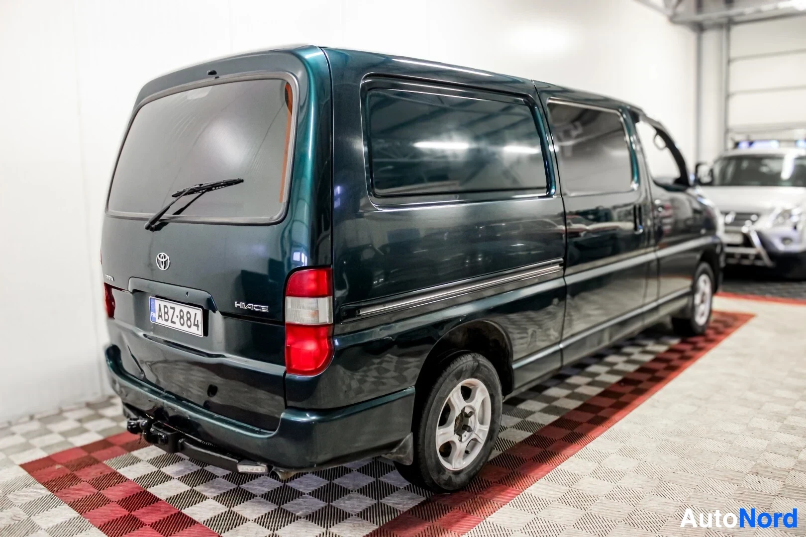 Takaviisto: Tummanvihreä pakettiauto, jossa on takaluukku auki ja vetokoukku, seisoo ruudullisella lattialla. Ajoneuvon takapuoli ja oikea kylki näkyvät, vasemmalla puolell