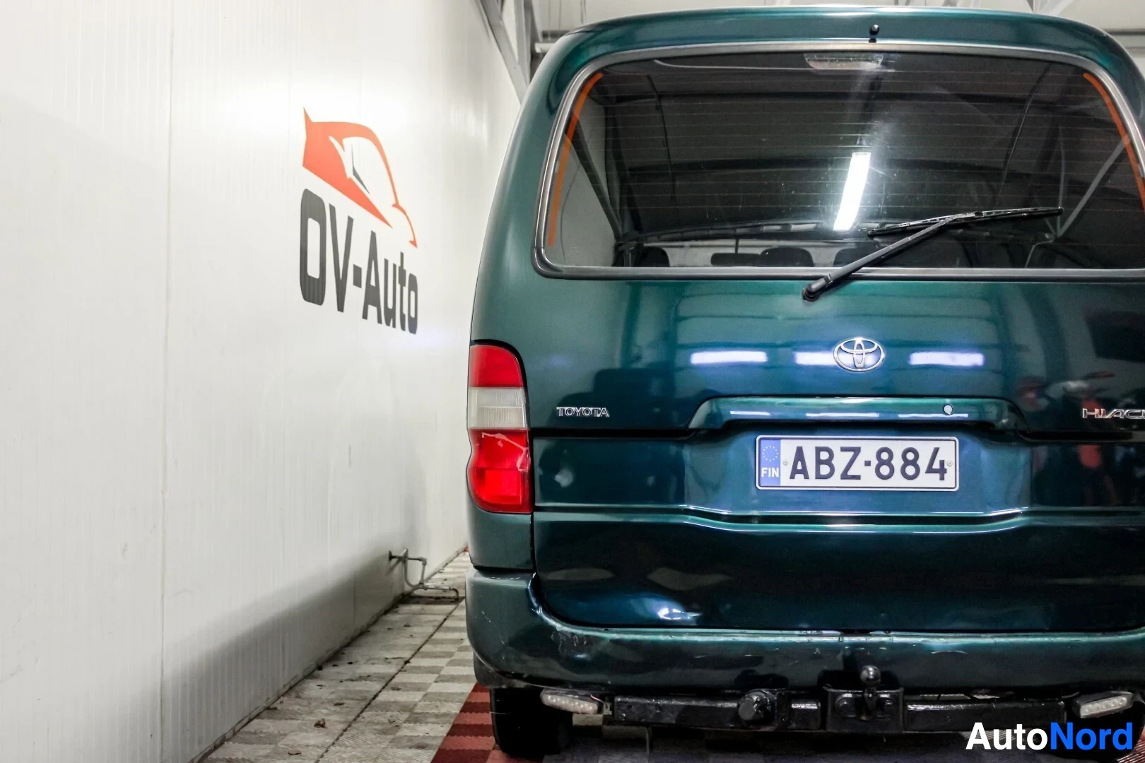 Takaa: Lähikuva tummanvihreän pakettiauton takaosasta, jossa näkyy takaluukku, takavalot ja rekisterikilpi. Valkoisessa seinässä takana lukee "OV-Auto".