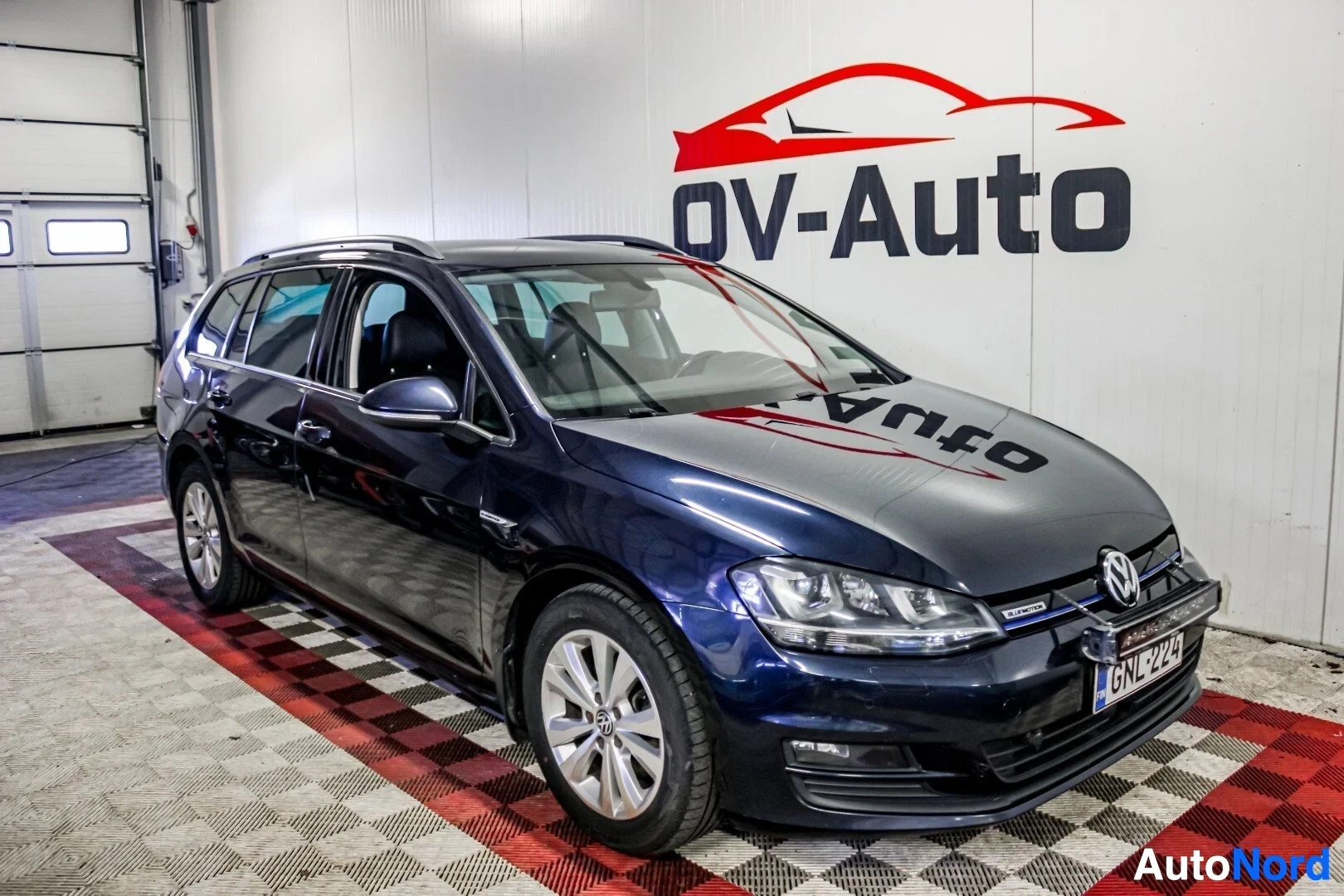 Etuviisto, nokka vasemmalle: Tummansininen farmariauto Volkswagen Golf Variant kuvattuna edestä oikealta puolelta sisätiloissa. Autossa on kattokaiteet ja kevytmetallivanteet. Reki