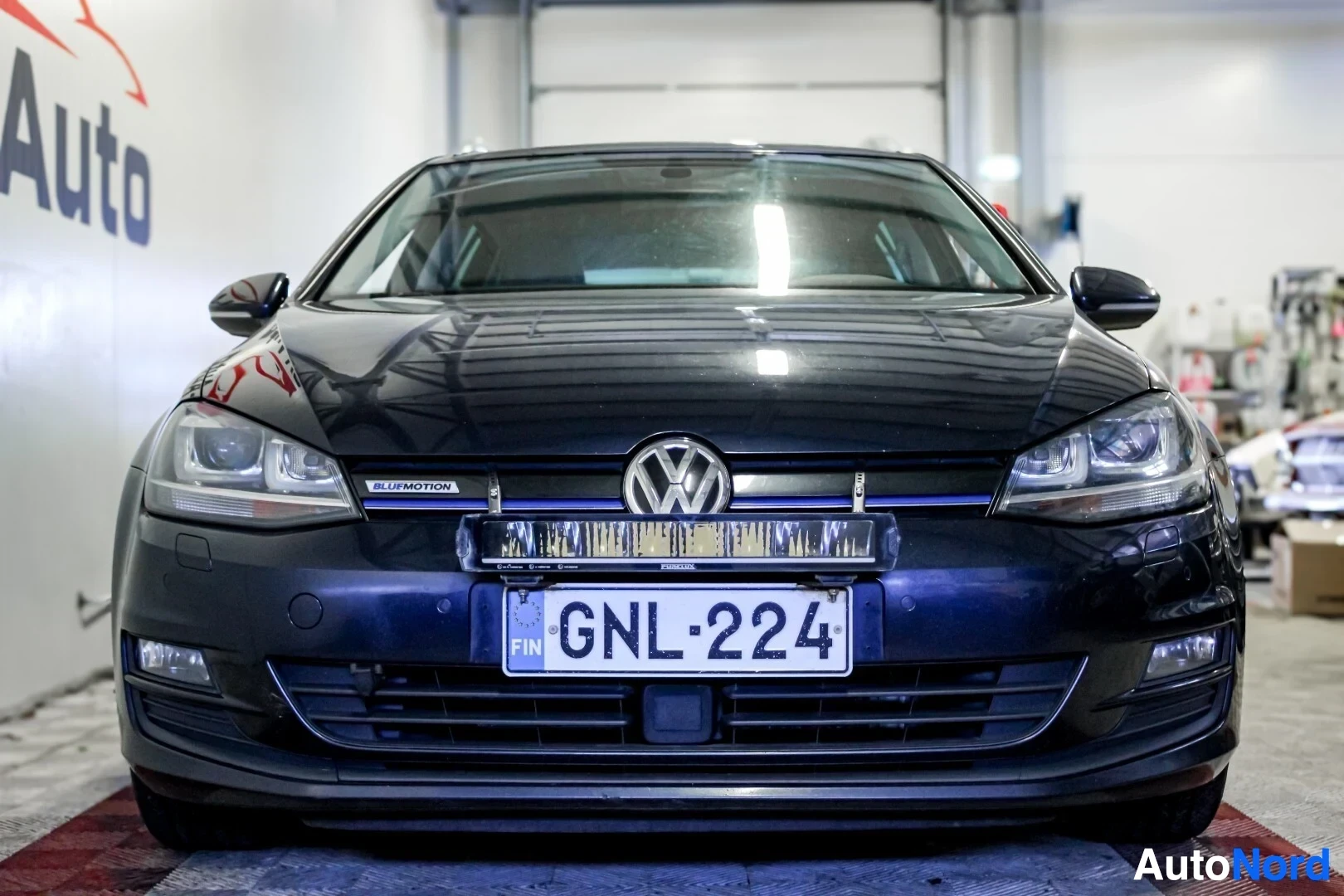 Edestä: Tummansinisen Volkswagen Golf Variant -farmariauton etuosa kuvattuna suoraan edestä. Eturekisterikilpi "" ja valopalkki ovat asennettuina puskuriin. Ajov