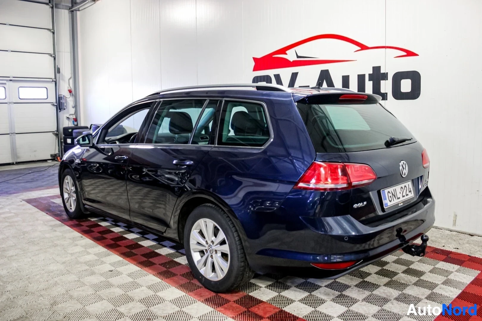 Takaviisto: Tummansininen farmariauto Volkswagen Golf Variant kuvattuna takaa oikealta puolelta sisätiloissa. Autossa on kattokaiteet ja vetokoukku. Rekisterikilpi "