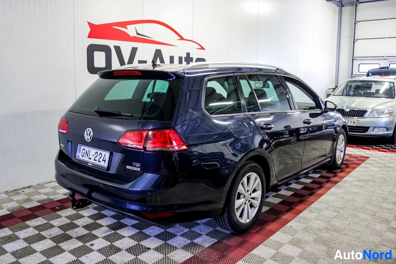 Takaviisto: Tummanvärisen farmariauton takaosa ja oikea kylki viistosti takaa kuvattuna autohallissa. Autossa on kattokaiteet ja vetokoukku.
